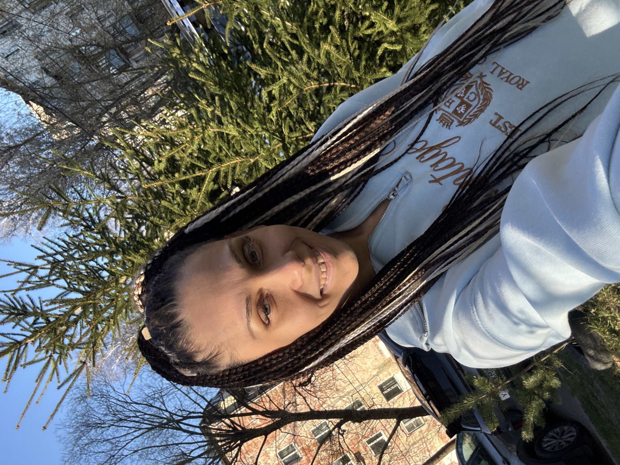 A smiling woman with long, two-tone braided hair (dark brown/black and light blonde/white) is captured in a bright, outdoor selfie taken in Rusanovo, Russia. She is looking directly at the camera with a joyful expression.
The woman is wearing a light blue zippered hooded sweatshirt (hoodie). On the front of her hoodie, there is a prominent golden-brown embroidered emblem resembling a crest or shield. Around this emblem, text is visible, including the word "ROYAL" at the top and "EST." (likely short for "Established") below it. Further down, there is a word in a flowing script font, which is partially obscured but appears to read "Vintage" or a similar word.
The setting appears to be a residential or urban area. In the background, there is a clear, bright blue sky. Lush green evergreen trees are prominent, suggesting a temperate climate. Bare deciduous trees, indicating late autumn, winter, or early spring, are also visible. Behind the trees, parts of multi-story buildings with windows are discernable, possibly constructed of brick or light-colored materials. A dark-colored car is partially visible parked on what appears to be a street or driveway in the lower part of the image. The bright sunlight suggests it is daytime.