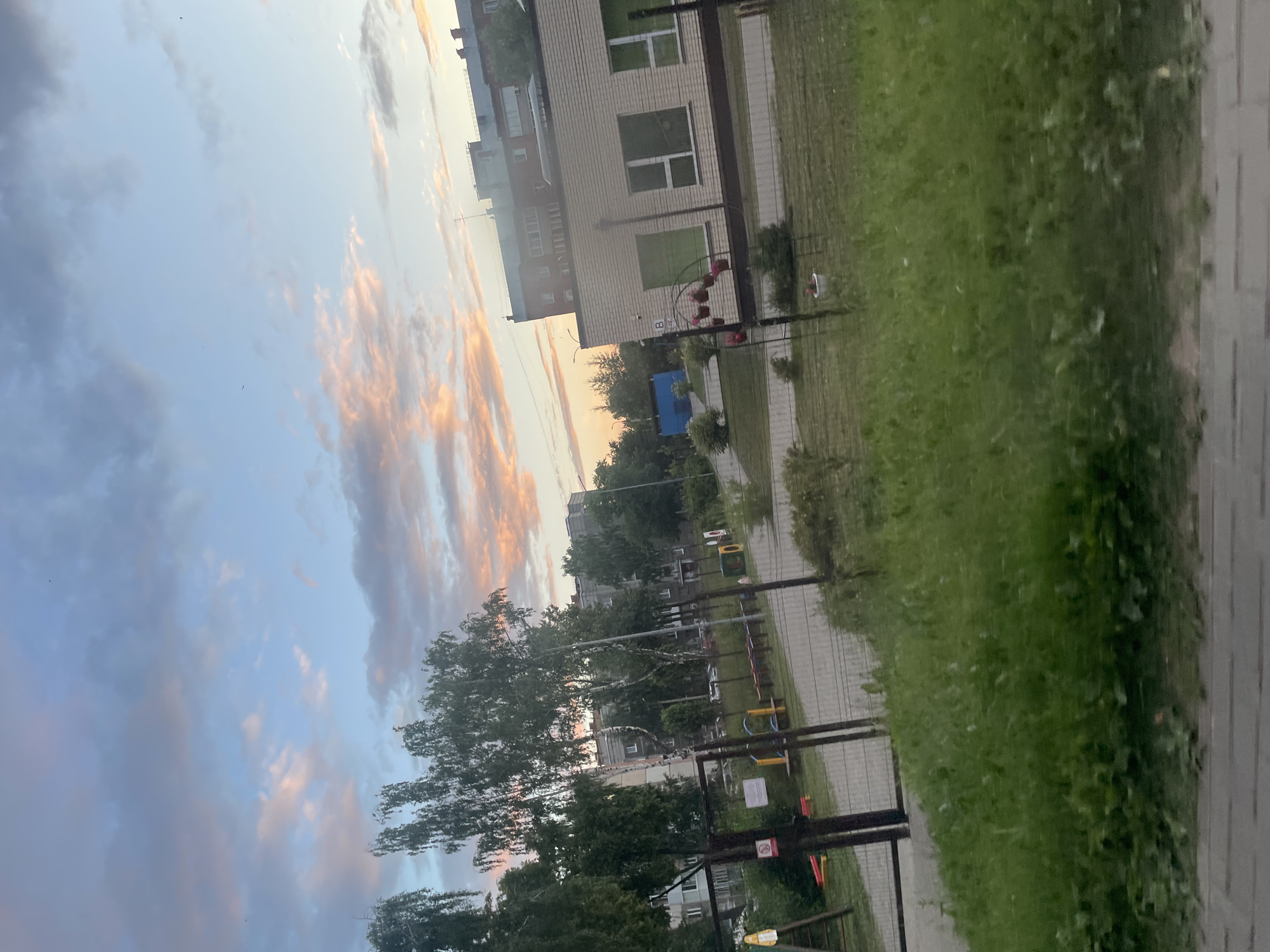 This media file depicts an outdoor scene with a sky filled with soft, colorful clouds in shades of pink, orange, and blue, suggesting it is either sunrise or sunset. The image is oriented vertically. In the foreground, there is a paved pathway bordered by a grassy area. Beyond the grass, a children's playground with colorful equipment, including swings and a slide, is visible. A metal fence separates the playground from the viewer. Further in the background, there are trees and residential buildings. One building, appearing to be a light-colored brick structure with multiple windows, stands out. There are also other apartment buildings and trees with lush green foliage. The overall impression is that of a peaceful urban or suburban setting during twilight. There is no discernible activity or people captured in the image, making it a landscape or cityscape shot. The weather appears calm, and the soft lighting from the sky creates a serene atmosphere. No visible text is present in the image. The location context suggests Votkinsk, Russia, which aligns with the architectural style of the buildings and the type of playground equipment often found in such locations.