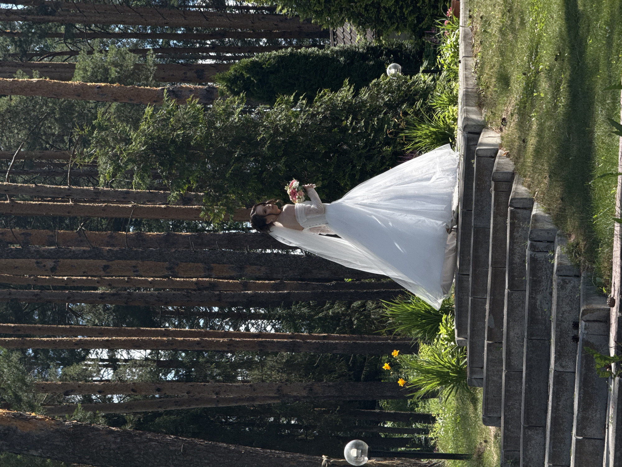 A bride, dressed in a white wedding gown and veil, stands on a set of concrete stairs in a wooded area. She is holding a bouquet of flowers. The trees are tall and thin, with a dense undergrowth of green foliage. The lighting suggests it is daytime, with sunlight filtering through the trees. The presence of pine cones on the stairs and the general ambiance of the scene suggest a natural, possibly park-like setting. The reference to Votkinsk, Russia, does not provide any specific visual cues within the image itself.
