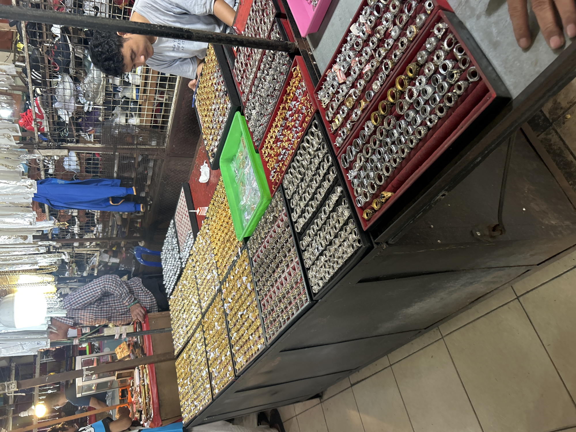 The image depicts a market stall in Denpasar, Indonesia, specializing in rings. In the foreground, a dark wooden display counter is laden with numerous trays of rings. These trays are arranged in rows and filled with a variety of rings, many appearing to be gold-toned and silver-toned, with some featuring dark stones or intricate designs. A man in a grey t-shirt is seated behind the counter, looking down at a tray of rings. To the left, a man in a checkered shirt is also present near the counter. In the background, shelves are stocked with various items, including what appear to be shoes and clothing. The overall environment suggests a bustling marketplace setting with goods displayed for sale.