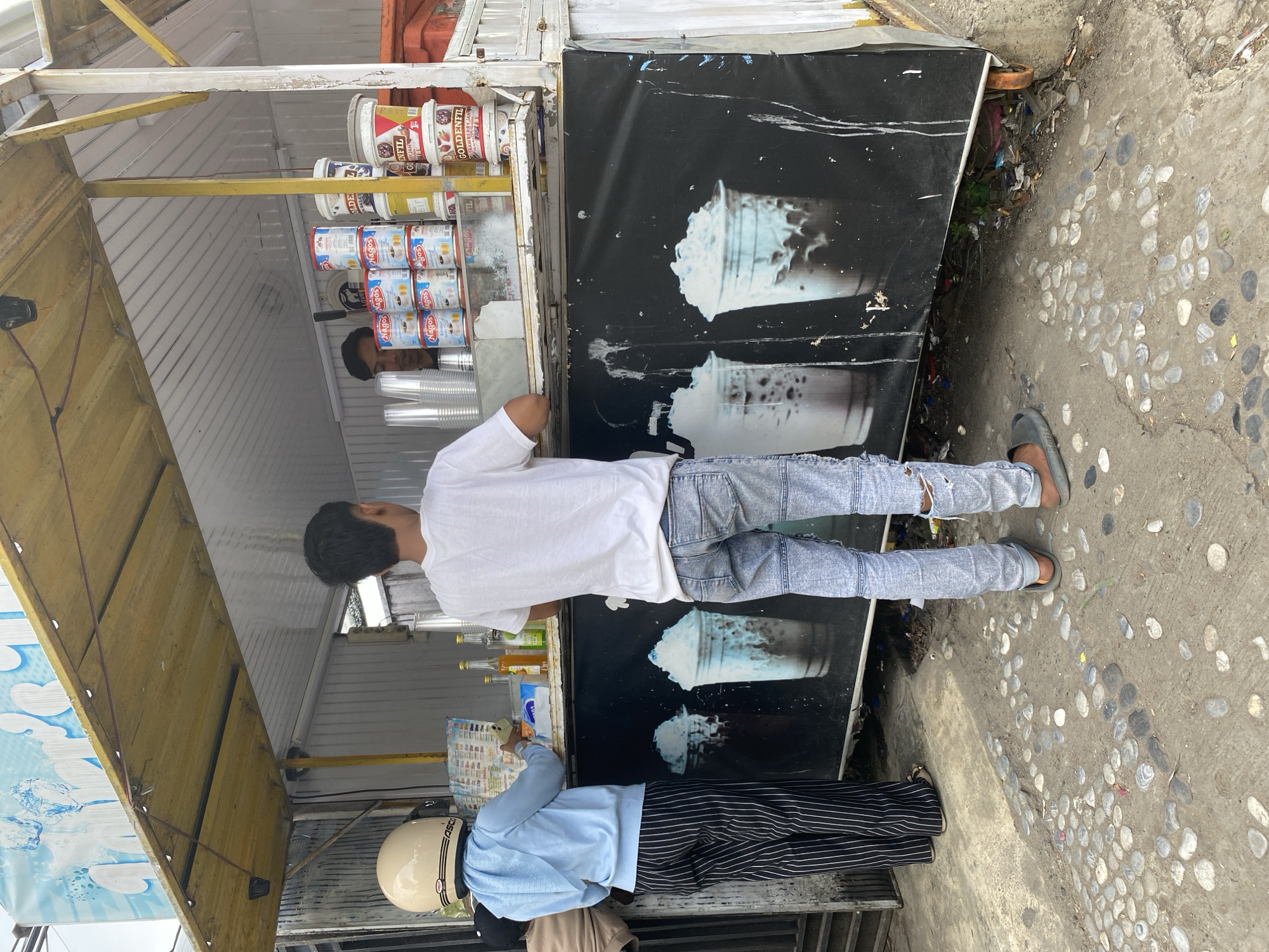 A street vendor's stall in Banda Aceh, Indonesia, is depicted, featuring two young individuals standing at the counter. The person on the left, facing the stall, wears a light blue hoodie, black and white striped pants, and a cream-colored helmet. They appear to be interacting with the vendor, who is partially visible behind the counter. The person on the right, facing away from the camera, wears a white t-shirt and distressed blue jeans, with gray sandals. The stall displays various beverage containers and products, including what appear to be cans of condensed milk and tubs of jam. The stall's exterior is decorated with images of iced drinks. The ground is paved with rough concrete and decorated with scattered stones. Debris is visible near the base of the stall. The overall scene captures a moment of commerce on a city street.