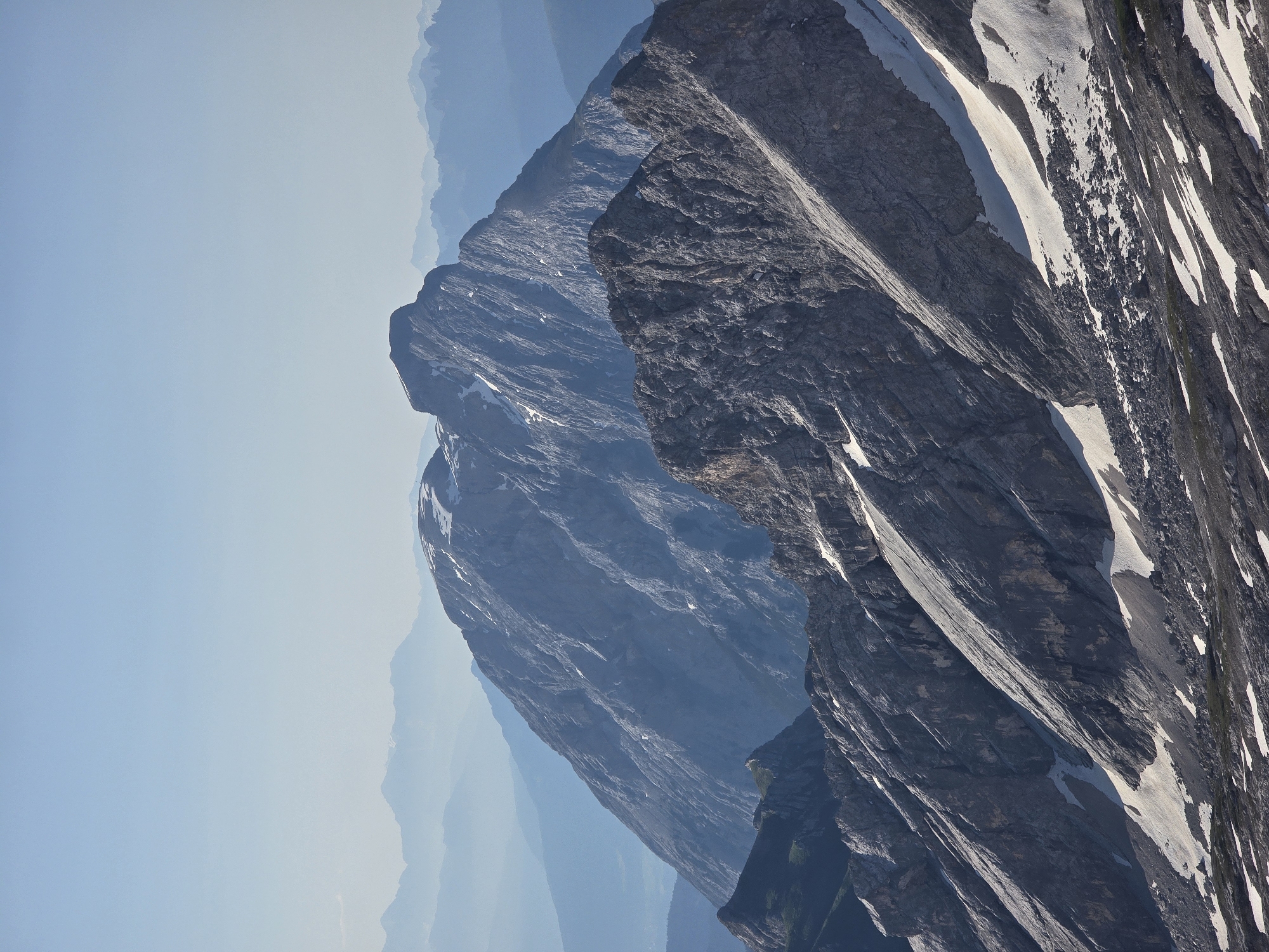 This is a vertical, long-shot, eye-level photograph of a mountain range. The sky is clear and light blue with a hazy gradient, giving it a soft, ethereal appearance. In the foreground, a rugged, rocky mountain slope descends diagonally from the left to the right. Patches of snow are scattered across the slope, contrasting with the dark gray rock. The texture of the rock is rough and craggy, with visible fissures and layers.
Further back, another prominent mountain peak rises, its surface also characterized by steep, rocky inclines and intermittent snow. This peak has a more defined, almost plateau-like summit. Behind this, a series of progressively lighter blue and more distant mountain ranges fade into the hazy horizon, creating a sense of immense depth and scale. The lighting suggests it is daytime, with the sun illuminating the mountains from the side, casting shadows that accentuate the rugged topography. The scene is devoid of human presence or any discernible man-made structures. The environment is exclusively natural, showcasing a majestic alpine landscape. This image likely depicts a scene in the Bavarian Alps, possibly near Garmisch-Partenkirchen, Germany, given the characteristic mountainous terrain and clear skies often found in that region.