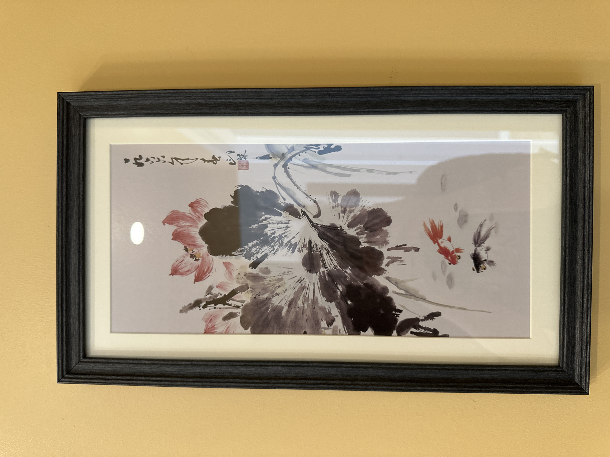A framed painting of lotus flowers and two goldfish hangs on a textured yellow wall. The painting is in a vertical orientation and is framed with a dark, textured border. The artwork depicts several large, dark lotus flowers with some pink petals visible, along with delicate blue tendrils. Two goldfish, one orange and white and the other dark gray, swim in the water below the flowers. There is some calligraphy in the upper right corner of the painting. The image appears to be taken indoors, possibly in a home or gallery setting, with a subtle reflection visible on the glass covering the painting. There is no visible indication of the city of Minsk, Belarus, or any specific location within it.