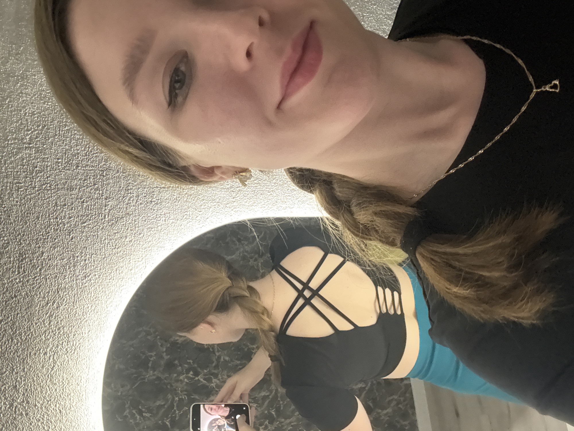 A woman is taking a selfie in a bathroom. She is holding her phone in front of a round mirror with a light behind it. The woman is wearing a black top with a crisscross back design and blue leggings. Her hair is in a braid. She has a gold necklace with a heart-shaped pendant. The wall behind the mirror has a textured surface. There is no indication of the location being Izhevsk, Russia.