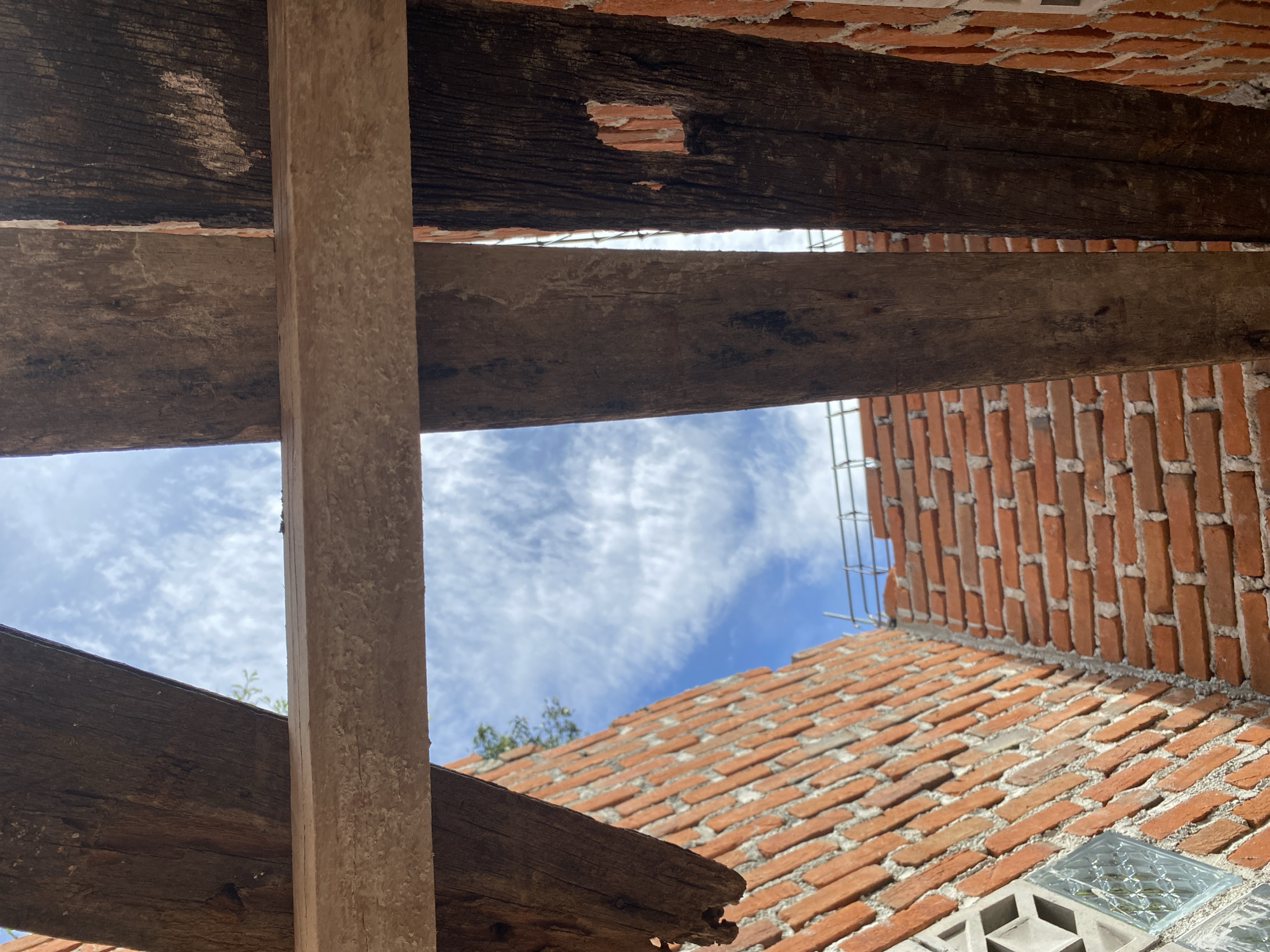 The image shows an unfinished brick building with wooden beams overhead against a blue sky with white clouds. The brickwork is in various stages of completion, with some sections appearing more solid and others still exposed. Two large, rough-hewn wooden beams run horizontally across the frame, obscuring parts of the sky and the building. The texture of the wood is visible, showing age and grain. In the background, scaffolding is visible, suggesting ongoing construction. The overall impression is one of a construction site with a view of the sky through an incomplete structure. The location is not specified in the image.