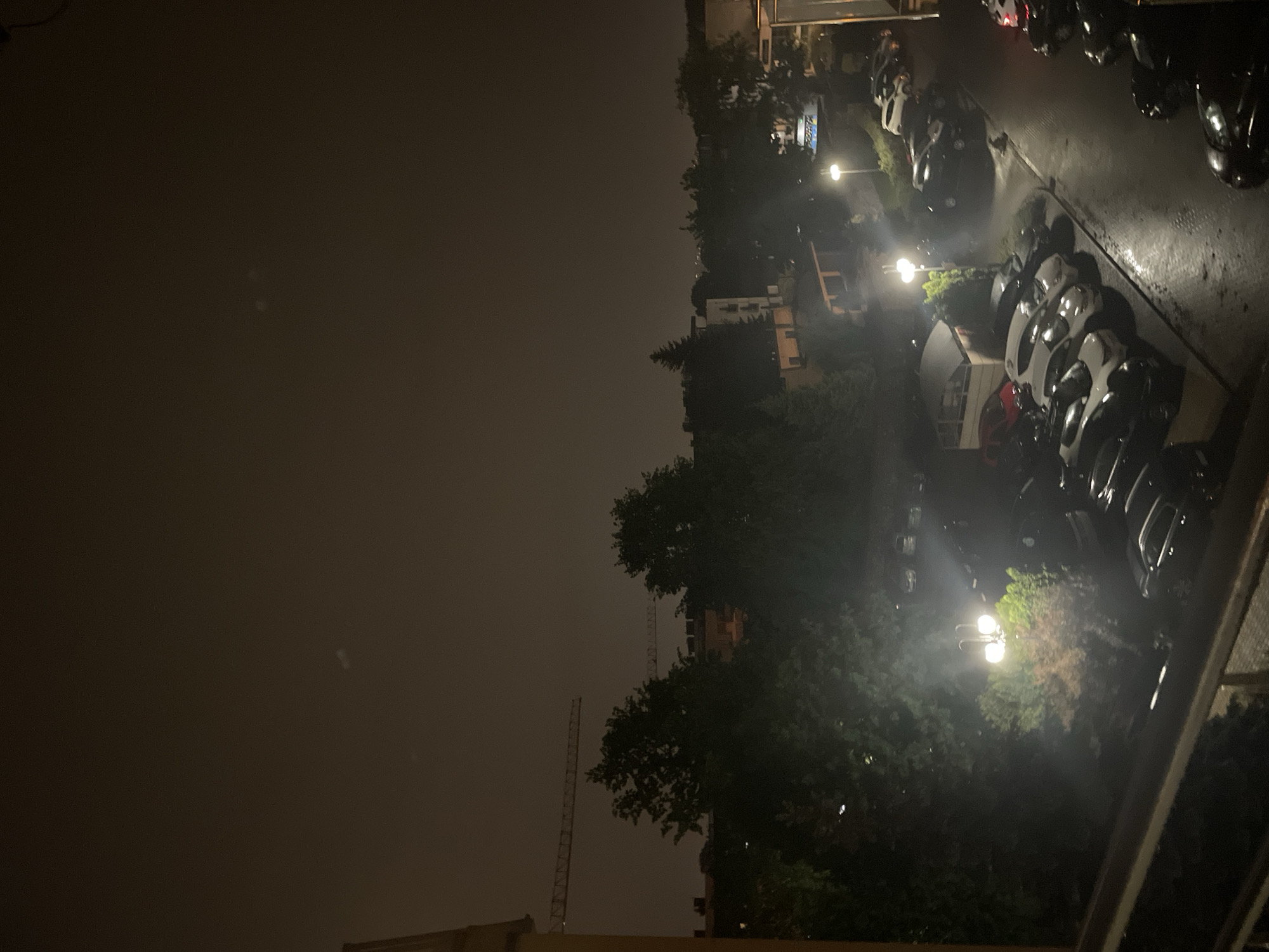 View from a balcony at night. The location is a parking lot next to a residential area with trees. Streetlights illuminate the cars. There is a construction crane in the background.