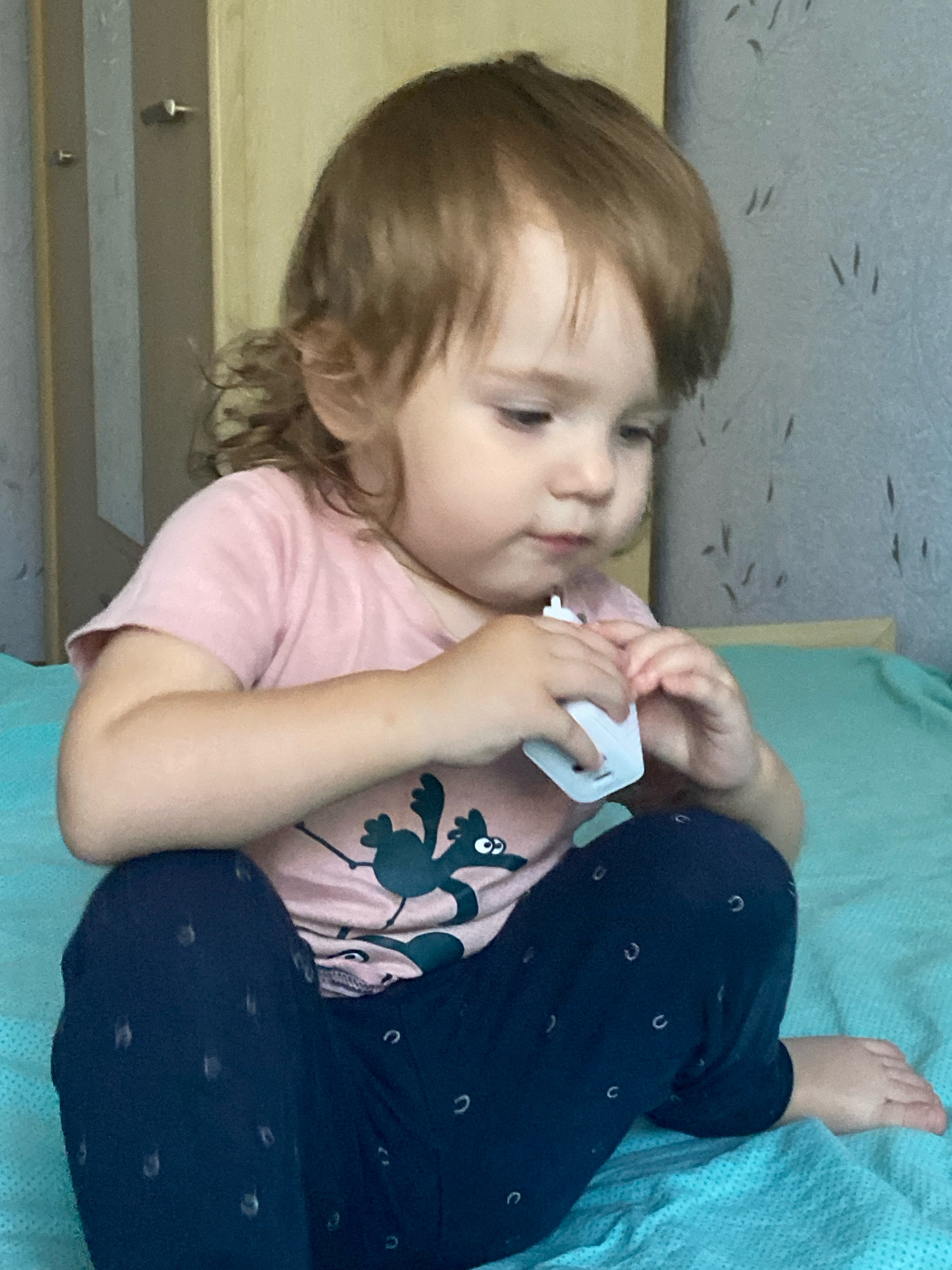 A young child with light brown hair and fair skin, wearing a pink t-shirt with a cartoon character and dark blue pants with a crescent moon pattern, sits on a turquoise blanket. The child is holding a small white object in their hands, appearing to be examining it. The background shows a wall with a light gray textured wallpaper and a wooden closet. The scene is indoors.