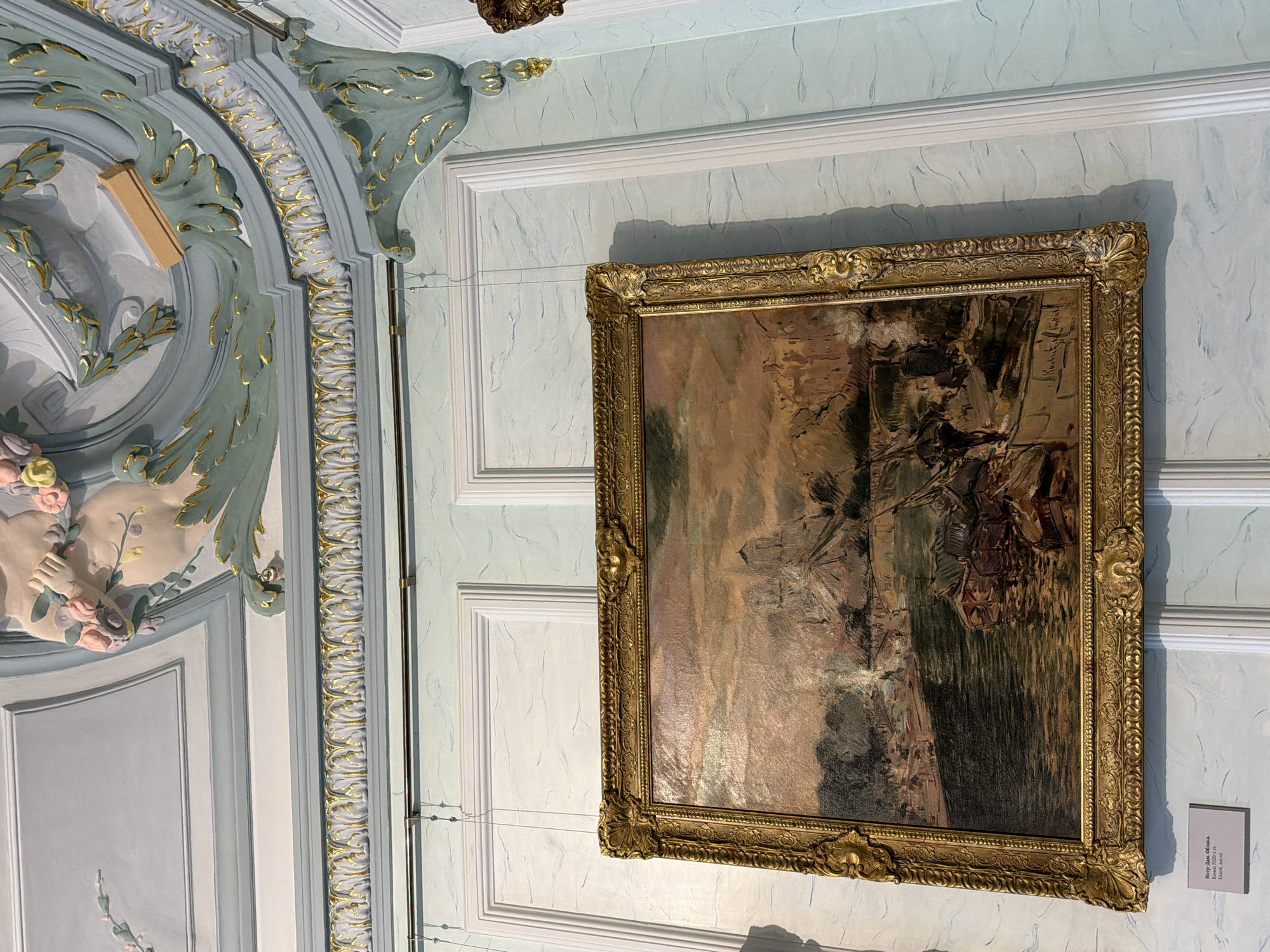 The image shows a painting displayed on a light blue wall with ornate white paneling. The painting is framed in an elaborate gold frame. Above the painting, the ceiling features decorative plasterwork in pale blue and gold. The painting itself depicts a busy river scene with numerous boats and buildings. The style of the painting suggests it might be an impressionist work. A small white label is placed below the painting. The location is identified as the Nizhny Novgorod State Art Museum, specifically the exhibition hall. The painting is titled "Notre Dame" and is dated to the late 19th - early 20th century, created with oil on canvas.