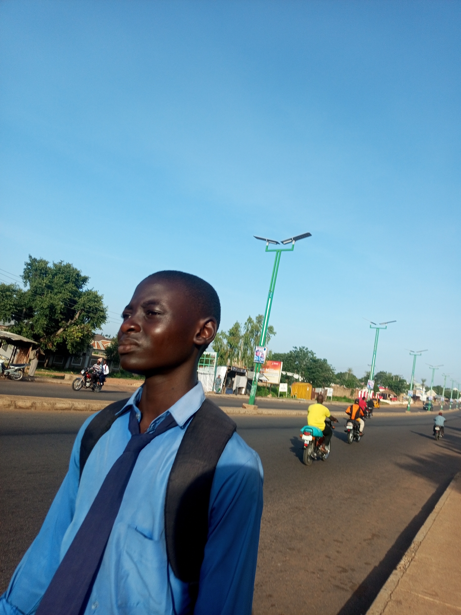 The image depicts a young boy wearing a blue shirt and a dark tie, with a dark backpack slung over his shoulders. He is standing on a street in Garko, Nigeria, looking to his left. In the background, a road is visible with several motorcycles and riders. The road is lined with trees and buildings, including some stalls. Modern streetlights with solar panels are also present along the road. The sky is clear and blue. The scene appears to be an everyday street view in the city.