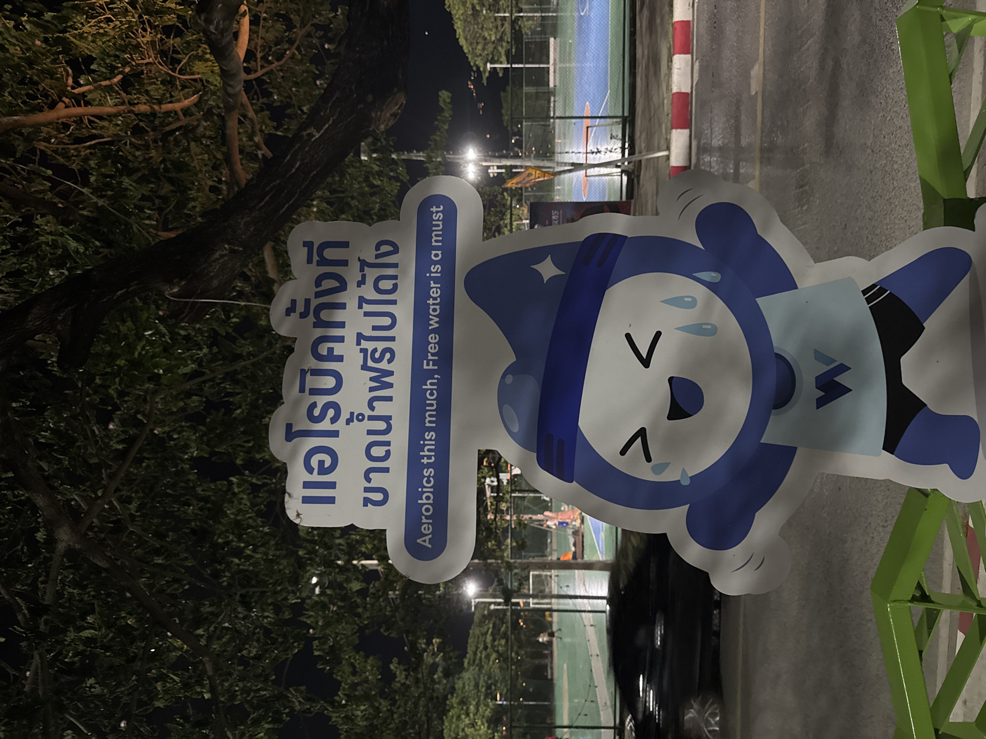 The image captures a nighttime scene in a public outdoor area, possibly a park or recreational facility, in the city of Ko Samui District, Thailand. A large, cartoonish cutout of a blue character in athletic wear, with sweat drops and a cheerful expression, is the central subject. The character is holding its hands up in a gesture of exertion or celebration. Above the character, a sign displays Thai text and the English phrase "Aerobics this much, Free water is a must." The background shows a basketball court with lighting, trees with dense foliage, and a street with a car. The overall ambiance suggests an event or facility promoting fitness and hydration.