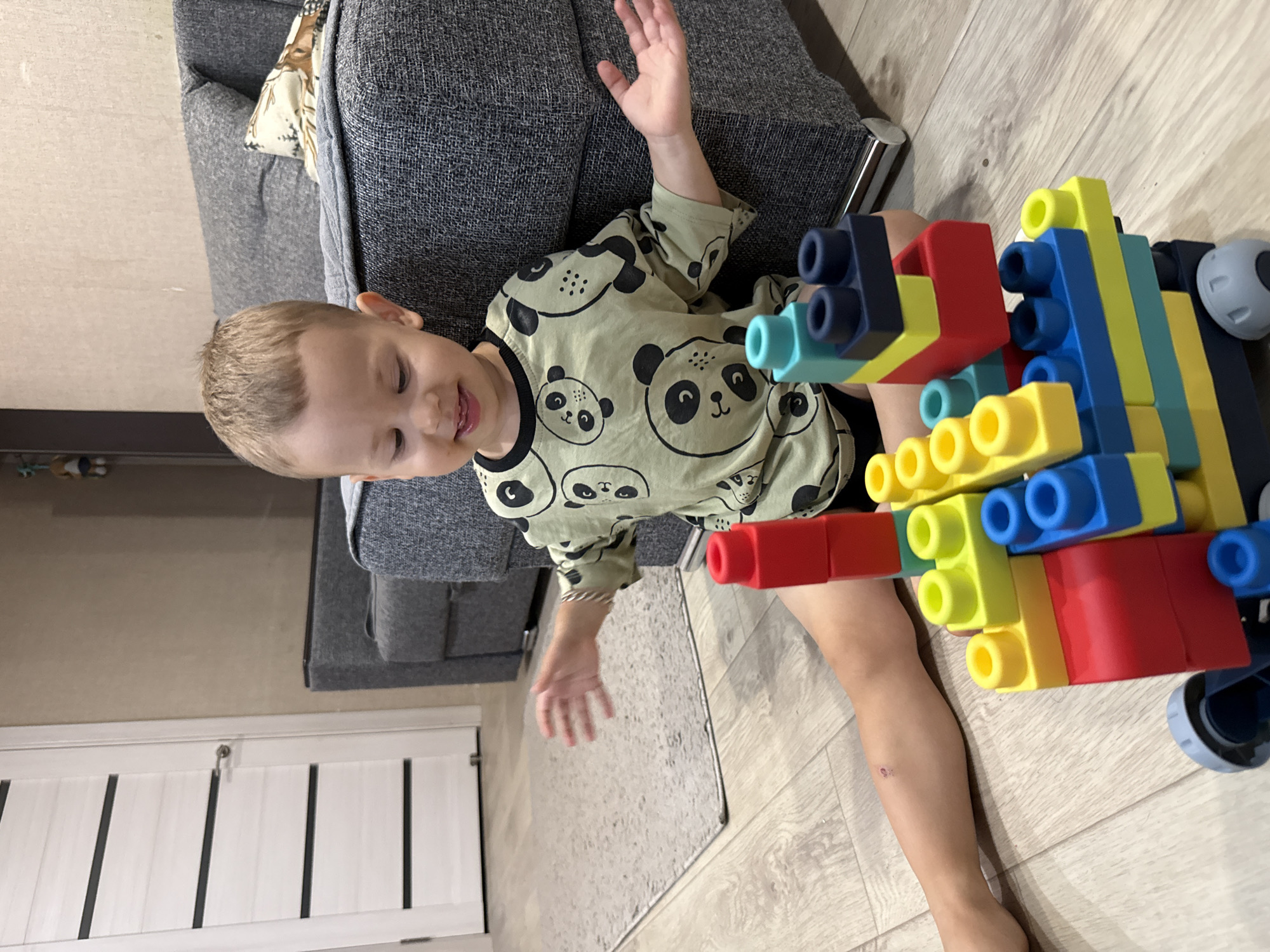 A young boy, possibly two to three years old, is sitting on a light wood floor, playing with colorful building blocks. He is wearing a light green long-sleeved shirt with a pattern of panda faces. The boy is looking down at the blocks, with his tongue slightly out, suggesting concentration. His right hand is extended outwards, and his left arm is bent at the elbow. Beside him, to his right, is a large grey sectional sofa. Behind the sofa, a cream-colored wall is visible. To the left of the boy is a light grey rug, and further left, a white door with horizontal dark stripes. The building blocks are stacked in a tower-like structure in front of the boy, featuring red, yellow, blue, and teal pieces, some with cylindrical connectors. The scene appears to be an indoor living space. The image does not provide any information or visual cues to place the location in Rusanovo, Russia.