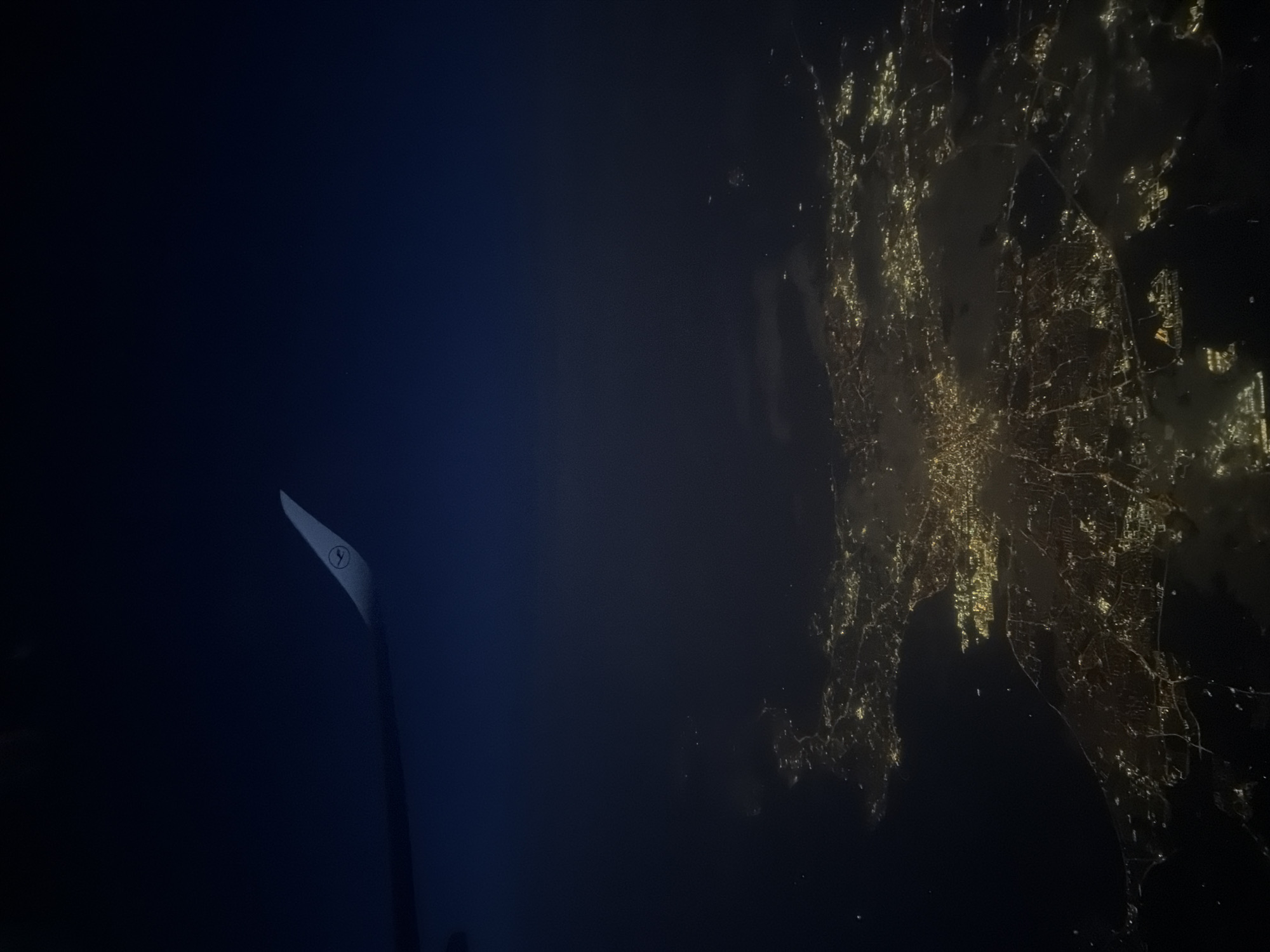 The image captures an aerial view at night, likely from an airplane. The right side of the frame displays a brightly lit urban area, presumably a city, with a dense network of streets and buildings illuminated by artificial lights. The left side of the frame is predominantly dark, representing the night sky and a portion of the airplane's wing. The wing has a logo on it. The city is not Eitting, Germany.