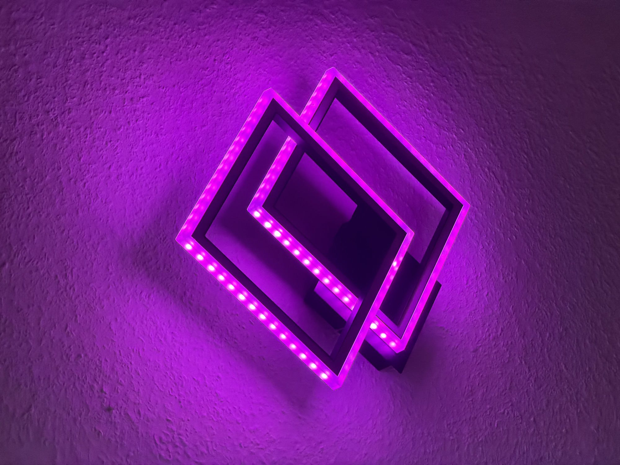 A close-up, angled shot shows a modern, geometric light fixture mounted on a textured, purple-lit wall. The fixture consists of two nested, square-shaped frames, each outlined with a strip of small, bright LED lights that emit a vibrant purple glow. The frames are slightly offset, creating a layered, three-dimensional effect. The wall behind the fixture is illuminated with the same purple light, highlighting its rough, uneven texture. The overall impression is one of contemporary design and atmospheric lighting. This image was taken in Dorsten, Germany.