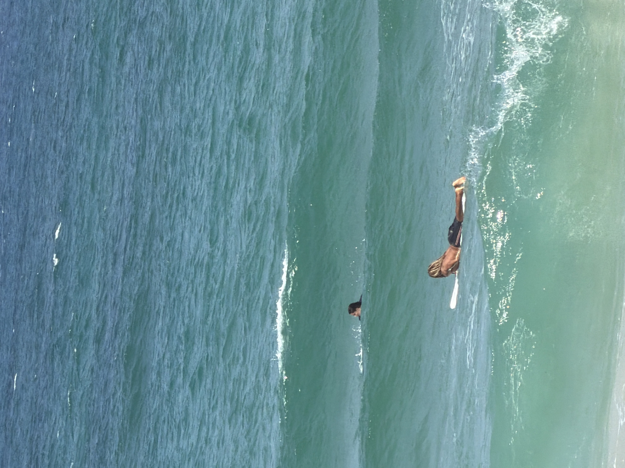 The image depicts an outdoor scene with two individuals in the ocean, presumed to be Tel Aviv, Israel, given the context provided.
In the foreground, a person is lying prone on a white surfboard, facing away from the camera. They are positioned on a wave, with their lower body submerged in the water and their legs extended backward. Their upper body is visible, and they appear to be tanned.
Further in the background, a second person is visible in the water, partially submerged. Only their head and shoulders are above the surface, and they are also facing away from the camera. Their hair is dark and wet.
The ocean is a dominant element in the image, characterized by turquoise-blue water with varying shades of green. There are gentle waves throughout the scene, with some white foam and spray visible, particularly around the surfboard and the second individual. The sky above is not clearly depicted. The overall impression is a sunny day at the beach with people engaged in water activities.