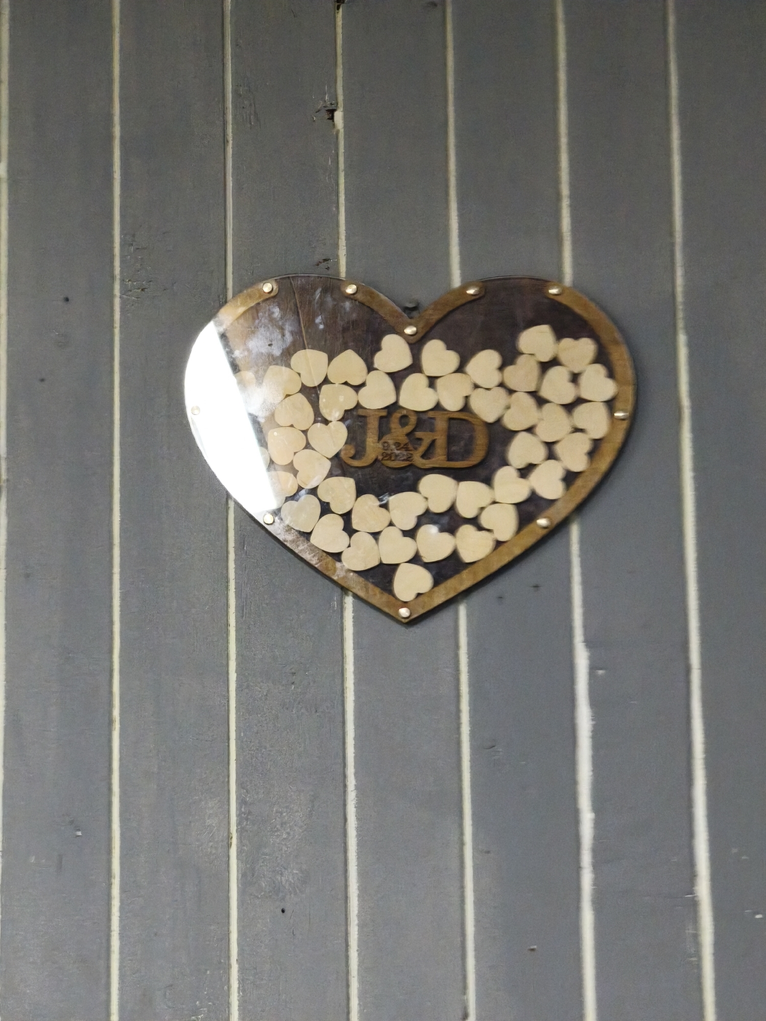 A wooden heart-shaped sign hangs on a gray wall with vertical white stripes. The sign features the letters "J&D" and the date "9.24 2022". Many small wooden hearts are attached to the sign. The sign appears to be a guest book or memorial of some kind. The location in Moscow, Russia, is not evident from the image.