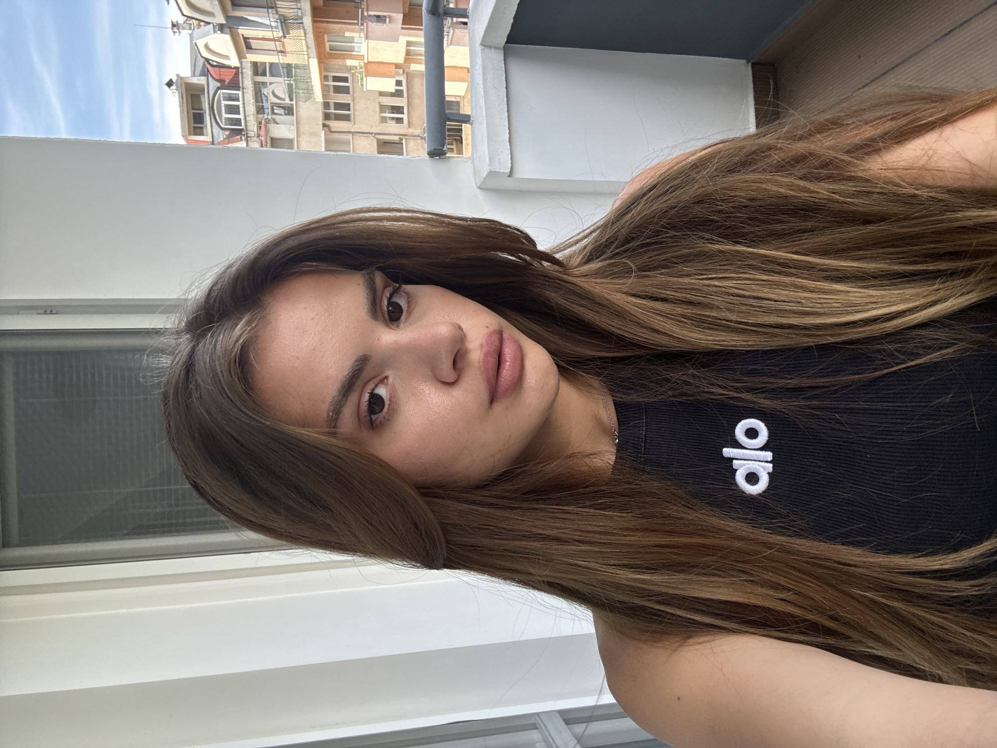 A young woman with long, flowing brown hair is featured in a close-up selfie, angled diagonally across the frame. She has a neutral expression and is looking directly at the camera. Her complexion is clear, and she has visible eyelashes and full lips with a natural-looking lip color. She wears a black ribbed tank top. Around her neck, a thin silver chain necklace is visible. On her top, near her right shoulder, the white "alo" logo is clearly visible.
The background suggests she is outdoors, possibly on a balcony or near a large window. To her left, a white window or door frame is visible. Beyond that, the upper parts of other buildings with multiple windows and balconies are seen under a clear, bright sky with sparse white clouds, indicating it is daytime. The architecture of the buildings in the background appears urban. Her long hair cascades around her shoulders and chest.