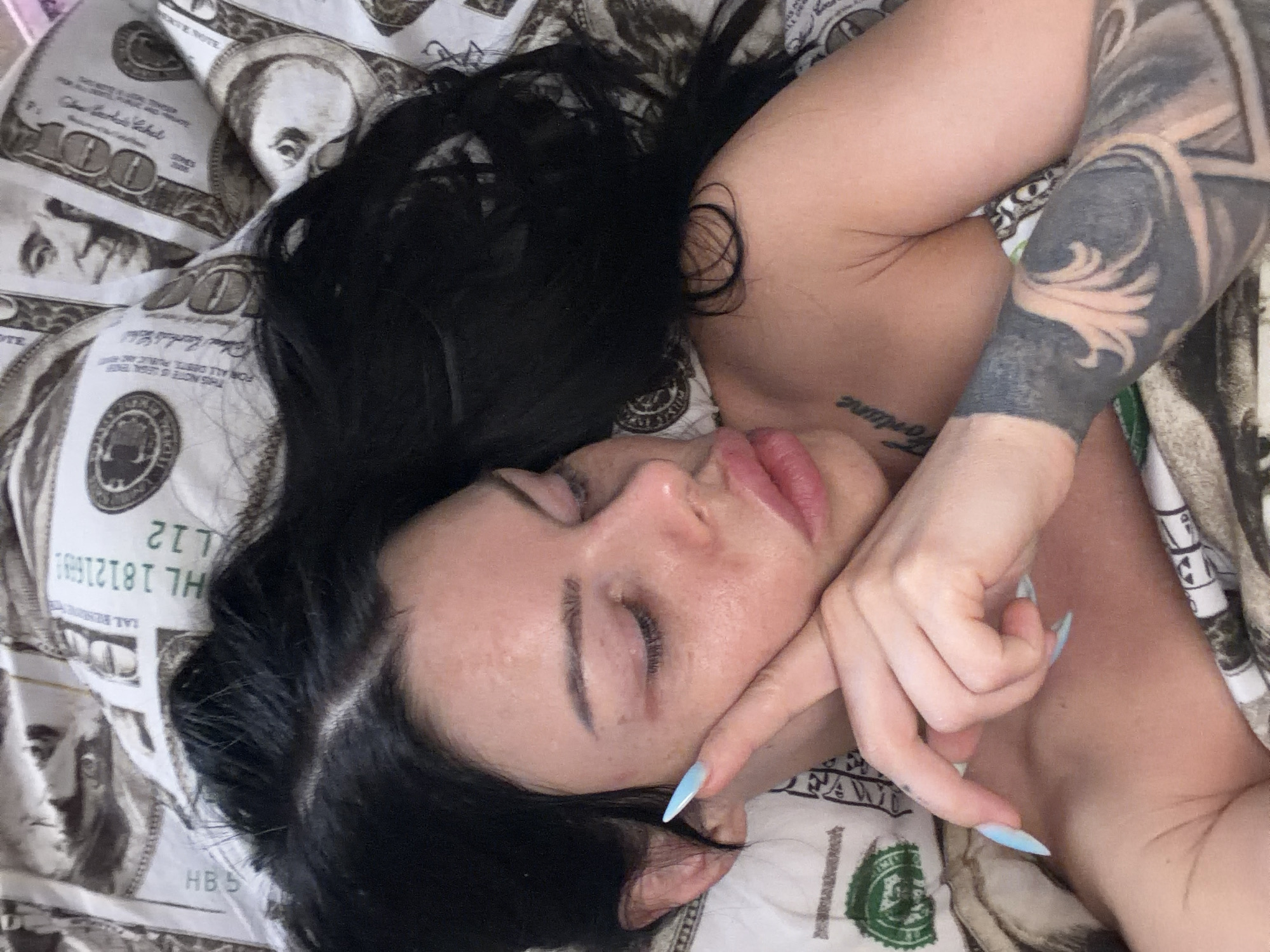 A close-up, overhead shot shows a woman with dark hair lying in bed. She is on her side, facing left, with her eyes closed and her right hand resting on her cheek. Her left hand is also near her face, with long, light blue nails. She has visible tattoos on her arm and chest. The bedding is patterned with images of US dollar bills. There is no indication of the city of Ufa, Russia in this image.