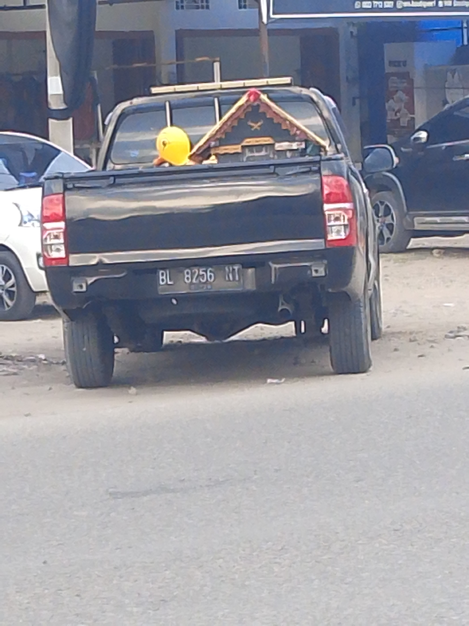 The image shows the rear of a black pickup truck parked on a paved road in Gampongpaya, Indonesia. The truck has a license plate that reads "BL 8256 NT". In the bed of the truck, a yellow object, possibly a balloon, and a small, ornate wooden structure with a pointed roof are visible. Another vehicle, a black SUV, is parked behind the pickup truck. The background shows buildings, suggesting an urban or semi-urban environment. The ground is a mix of asphalt and dirt.