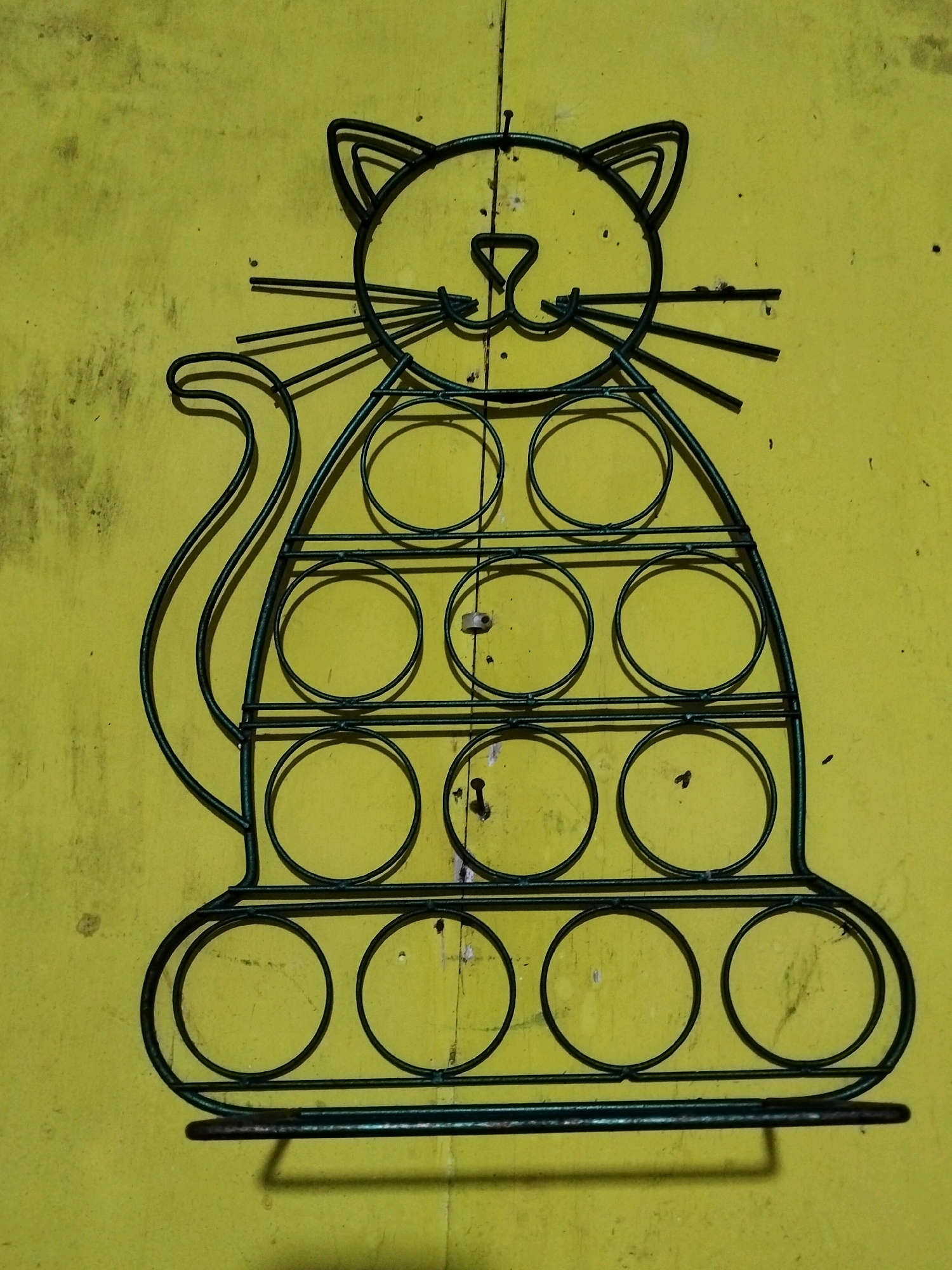 A green metal wire sculpture of a cat hangs on a weathered, yellow-green wall. The sculpture is stylized, with a round head featuring triangular ears, a small nose, and whiskers. Its body is also rounded and filled with a grid of ten circular openings arranged in three rows. A curved tail extends from the left side of the cat's body. The sculpture appears to be a functional item, possibly a holder for small items. The wall behind it shows signs of age and wear, with scuffs and darker patches, suggesting it is an outdoor or utility space. The image is taken from a slightly elevated angle, with a shadow cast by the sculpture on the lower right. The location is identified as Teluk Pambang, Indonesia.