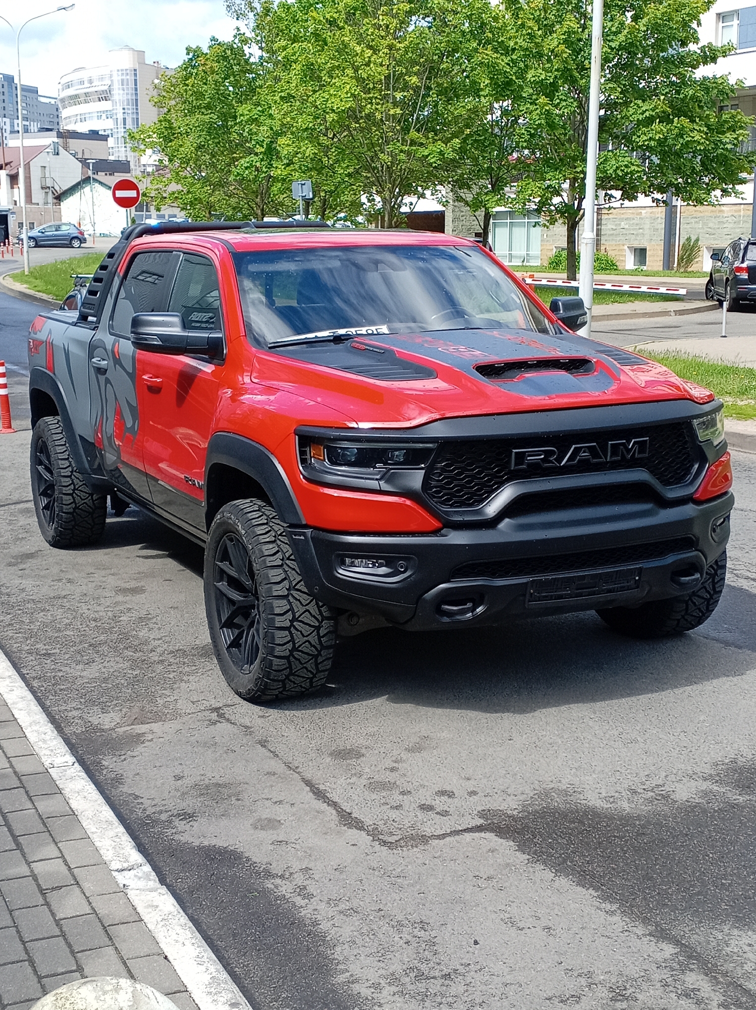 A red and gray RAM TRX pickup truck is parked on a wet asphalt surface next to a brick sidewalk, with a painted white line separating the two. The truck features black accents on the hood, grille, wheels, and side details. A red and white "no entry" traffic sign is visible in the background, along with a row of orange traffic cones. Buildings and trees are also visible in the background. The location appears to be an urban street, although the city and region are not specified.