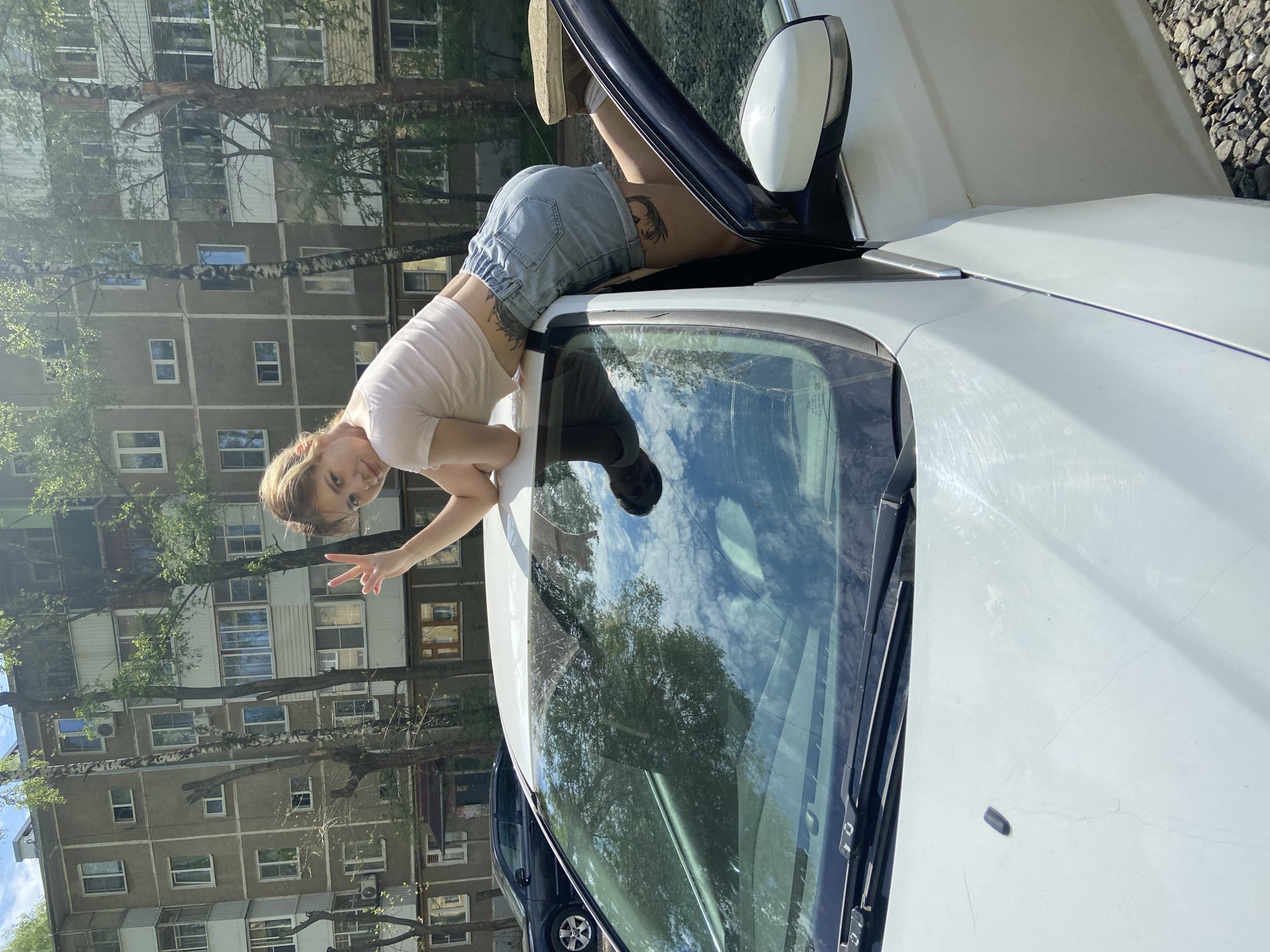 A young woman in a light-colored t-shirt and jean shorts is leaning over the hood of a white car with a partially open driver-side door. She is facing the camera, giving a peace sign with her right hand. Her feet are resting inside the open door. The car is parked in front of a large, multi-story apartment building with many windows. Trees are visible in the foreground and background. The reflection of trees and the sky is visible in the windshield. The location appears to be Ryazan, Russia, based on the architectural style of the building. The ground is partly covered with gravel. Another car, darker in color, is partially visible in the background.