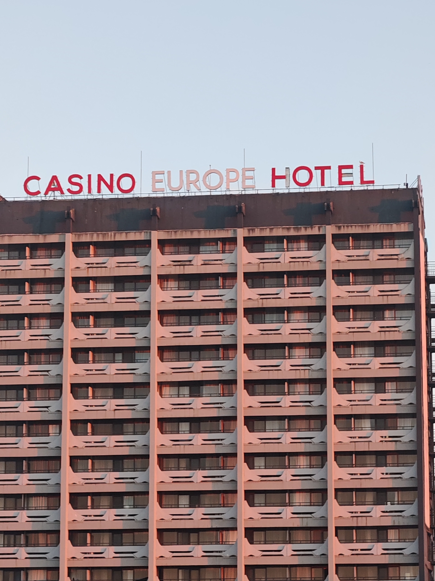 The image captures a high-angle view of the Casino Europe Hotel in Nesebar, Bulgaria. The hotel is a high-rise structure with a repetitive facade of windows and balconies. The hotel's name is displayed on the rooftop in red and white block letters. The building's exterior exhibits signs of weathering. A fire escape is visible on the right side of the building. The sky is pale blue.