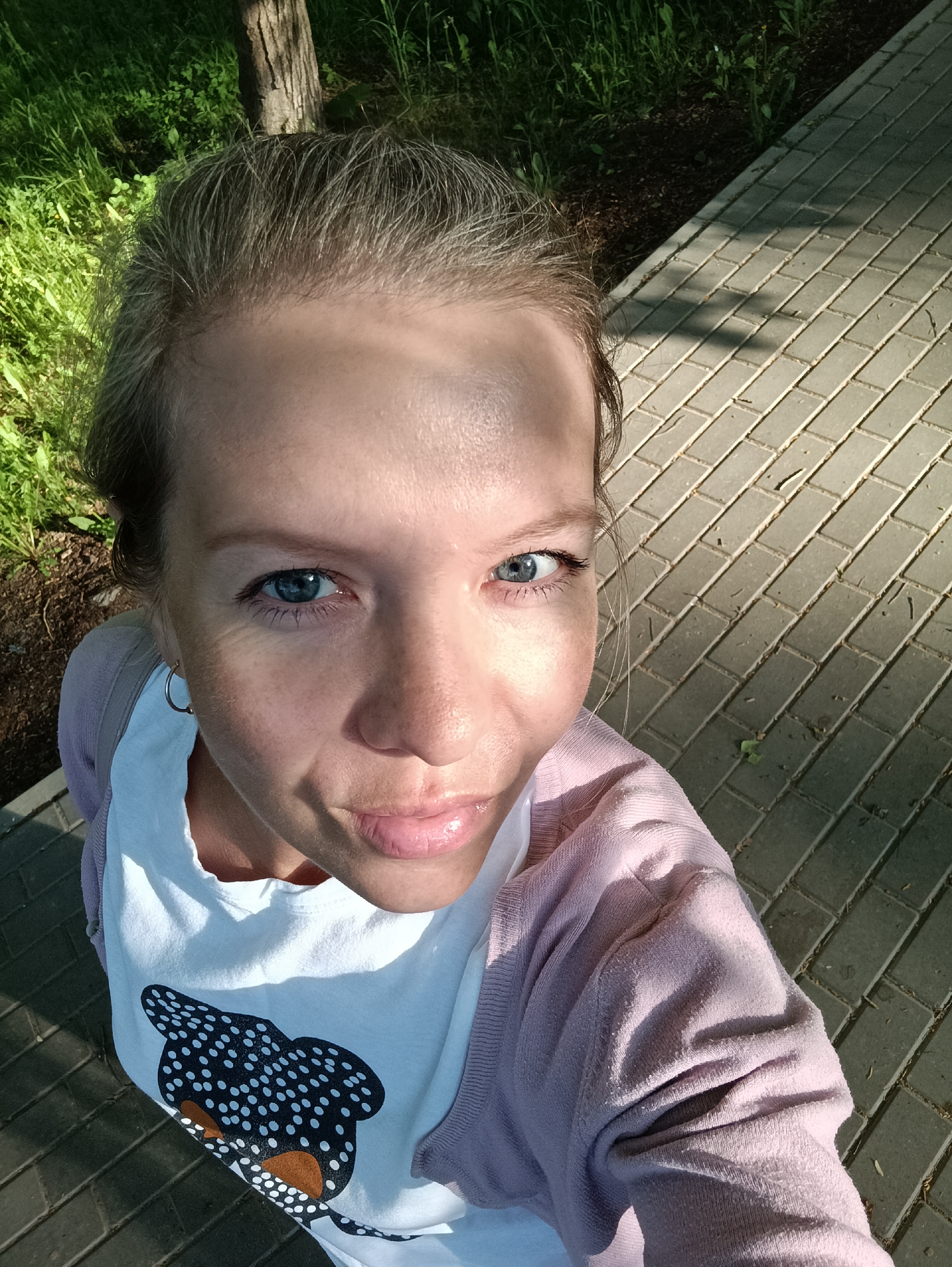 A high-angle, close-up self-portrait shows a woman with blue eyes and light skin, standing on a brick-paved walkway, partially in shadow. She is wearing a white t-shirt with a black and brown design resembling an animal and a light purple cardigan. Behind her are patches of grass and a tree trunk. The location may be in the city of Chaikovsky, Russia.