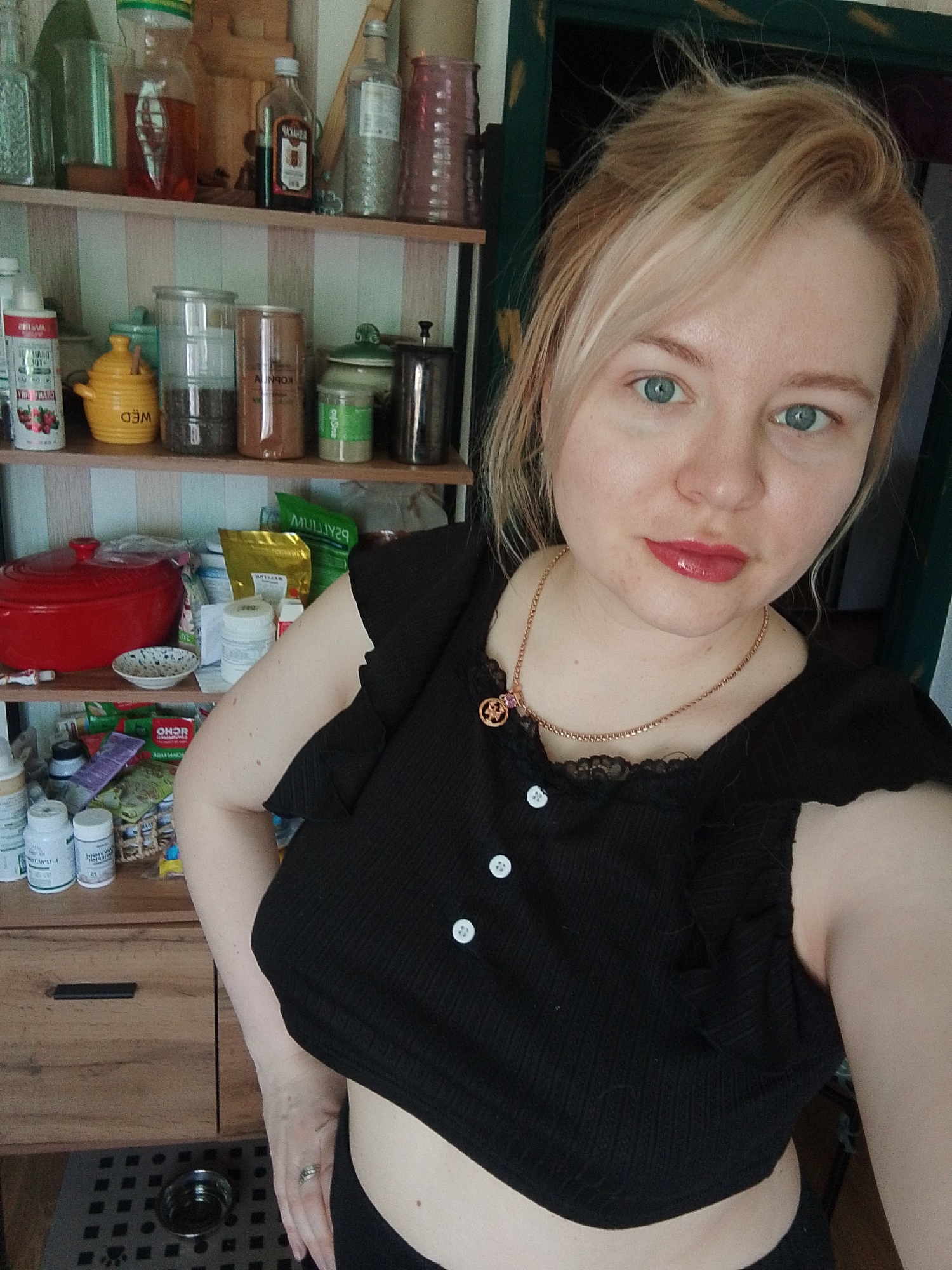 A light-skinned female with blonde hair, styled loosely with some strands on her forehead, is depicted from the mid-torso upwards, taking a selfie indoors. Her blue-green eyes are directed towards the camera, and her lips are painted with a light red/pink glossy substance. She wears a black ribbed crop top with ruffled short sleeves and three visible white buttons down the front. A golden-colored chain necklace with a small circular pendant, featuring a possible star motif and a pink jewel, is around her neck. A silver-colored ring is visible on her left ring finger.
The background reveals an indoor setting, consistent with a kitchen or pantry. Light-colored wallpaper with subtle vertical stripes covers the wall. Two wooden shelves are visible behind the subject. The top shelf holds several glass bottles, including one with amber liquid labeled "КАГОРЪ" (Kagor), another with light liquid and herbs, and a ribbed pink glass jar. The middle shelf displays various items: a yellow ceramic pot with "WED" (likely "DEW" reversed) inscribed, clear jars with dark contents, a brown cylindrical container (possibly labeled "КОРИЦА" - Cinnamon), a green lidded ceramic jar, a metal grinder, a red ceramic pot with a lid, a small white patterned bowl, and several plastic bags, one of which partially shows text resembling "PSYLIUM." The lower portion of the image reveals a wooden cabinet or drawer unit with a dark handle, and on the floor, a dark gray square-patterned mat with a small silver metal pet bowl resting on it. Several white plastic bottles, potentially supplements, are visible on a surface to the left. A dark green door frame is partially visible in the upper right.
The specific location within the city of Izhevsk, Russia, cannot be determined from the visual information provided in the image.