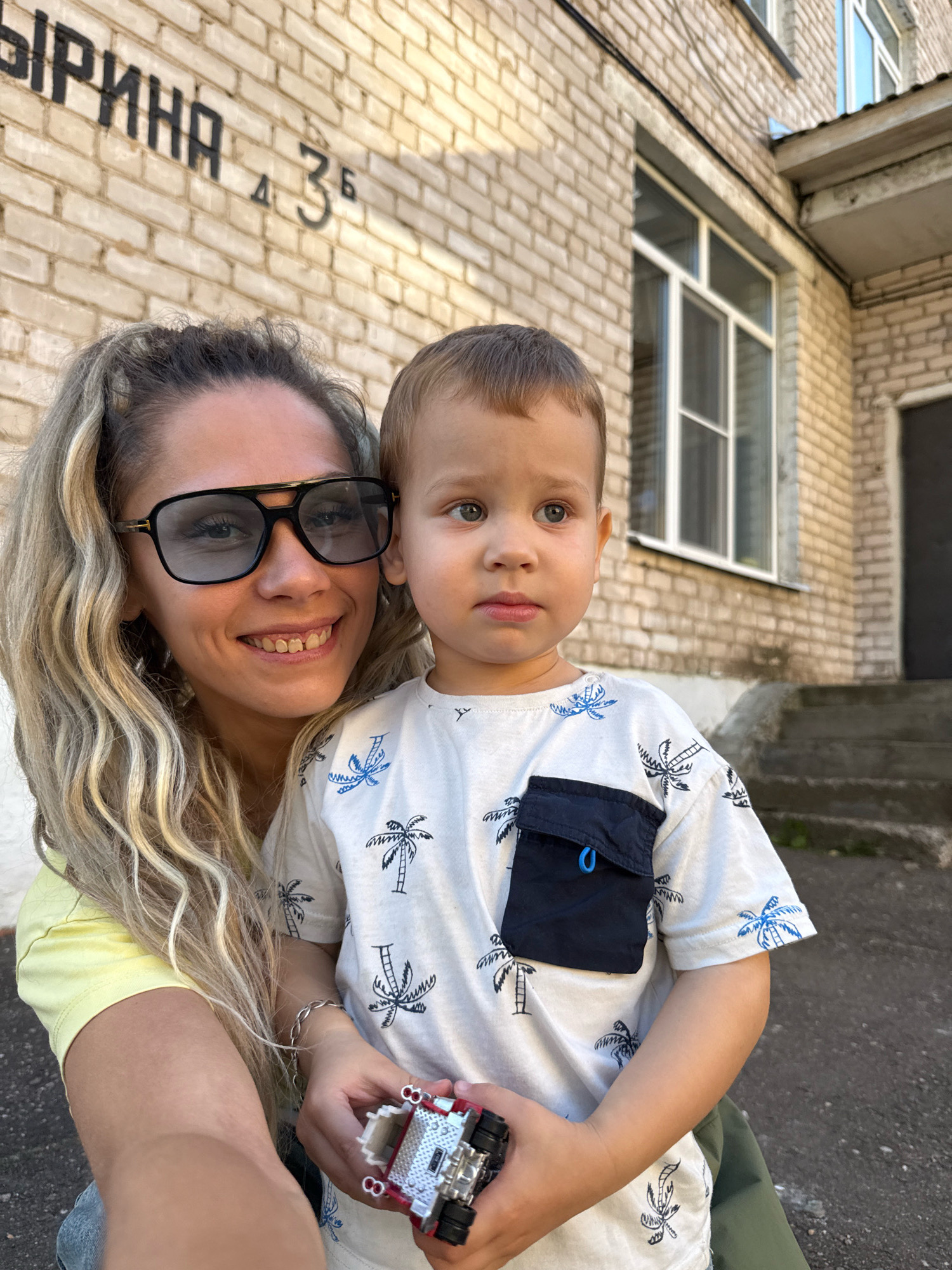 In this outdoor, eye-level medium shot, a young boy and a woman are pictured close together. The woman, on the left, is smiling broadly and wearing large, dark sunglasses and a light yellow top. Her blonde hair is styled loosely, with some strands framing her face. She is holding a small, red and silver toy vehicle in her right hand, which is positioned in front of the boy. The boy, on the right, looks directly at the camera with a neutral expression. He has light brown hair and is wearing a white t-shirt with a pattern of blue palm trees and a dark blue pocket on the chest. He is looking slightly off to the side, and his right arm is held across his body, where he is holding the toy vehicle with the woman.
The background consists of a light-colored brick building. Above and to the left of the subjects, black lettering is visible on the bricks, which reads "ВЫРИНА" followed by "д 35". The building has windows and a dark door visible to the right. Concrete steps lead up to the building on the right side of the frame. The lighting suggests it is daytime, possibly late afternoon due to the warm tones. The overall mood appears casual and affectionate.