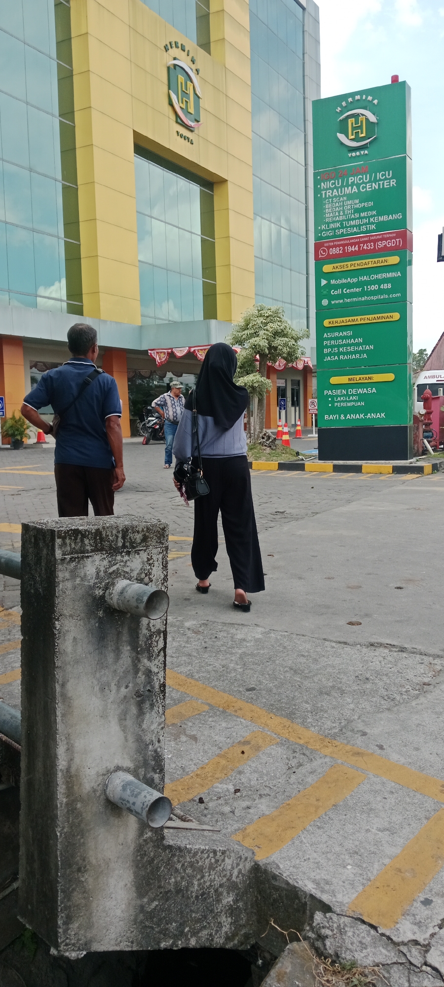 The image shows the exterior of a building identified as "HERMINA YOGYA" with a prominent yellow facade and glass windows. A green signpost stands to the right of the building, displaying information in Indonesian. In the foreground, a concrete structure with a pipe is visible. Two individuals are walking away from the camera towards the building: a man in a blue shirt and brown pants, and a woman wearing a black hijab and black pants. Another man is standing in the background near a motorcycle, looking towards the building. The scene appears to be a public outdoor area, possibly a parking lot or walkway, with yellow and black striped lines painted on the concrete ground. The environment suggests an urban setting in Maguwoharjo, Indonesia, likely in front of a hospital or medical facility given the signage.