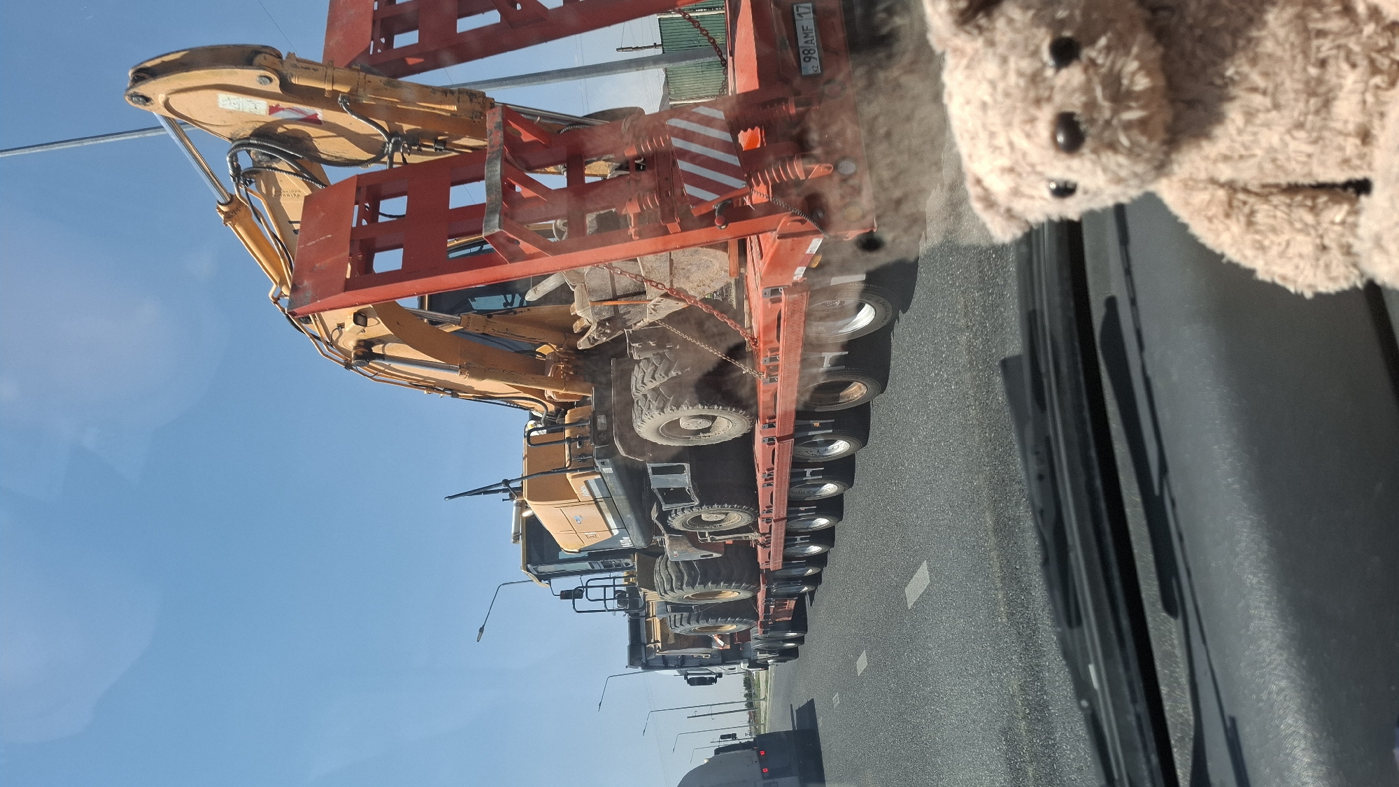 On a clear day in Shymkent, Kazakhstan, a flatbed truck transports two large yellow excavators along a highway. The truck, with its multiple axles, carries the heavy machinery, which are secured to the trailer. The excavator in the foreground is partially visible, its bright orange arm extending upwards. The road is asphalt, with white lane markings. In the foreground, a plush teddy bear is positioned on the dashboard of a vehicle, seemingly observing the scene. The sky is a bright, cloudless blue.