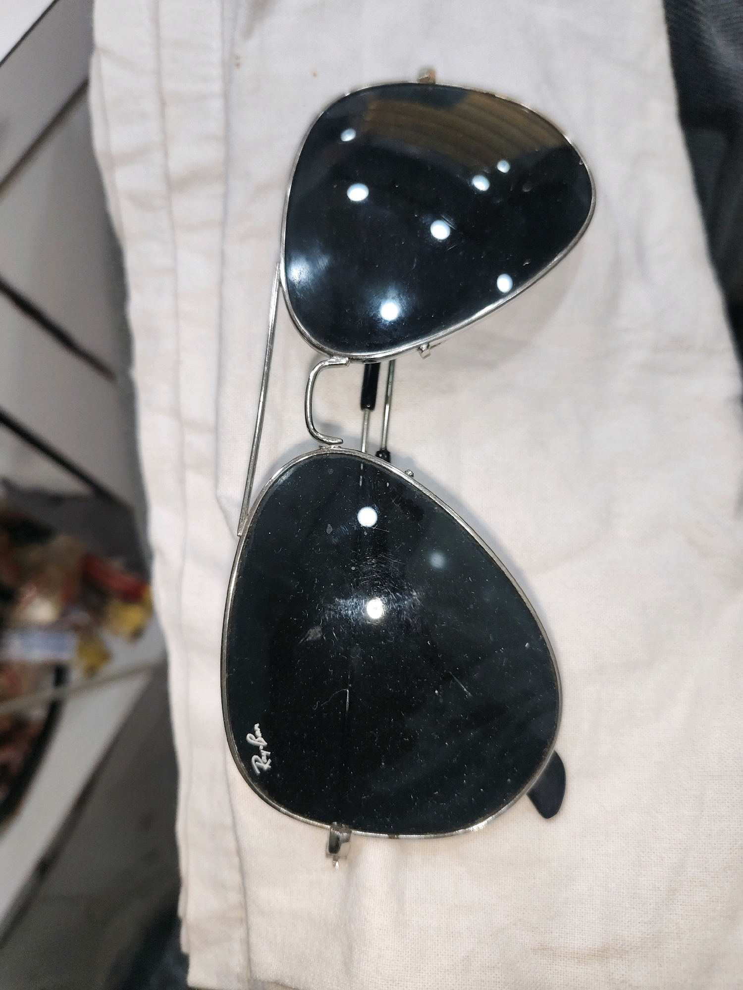 A pair of aviator sunglasses with black lenses and silver frames is resting on a white fabric surface. The sunglasses are positioned at an angle, with the lenses reflecting white lights. The fabric appears to be a shirt or cloth, with visible texture and some subtle creases. In the background, out of focus, is a glimpse of what appears to be a market or shop setting with shelves and items on them. The bottom left lens of the sunglasses shows a visible "Ray-Ban" logo etched onto it.