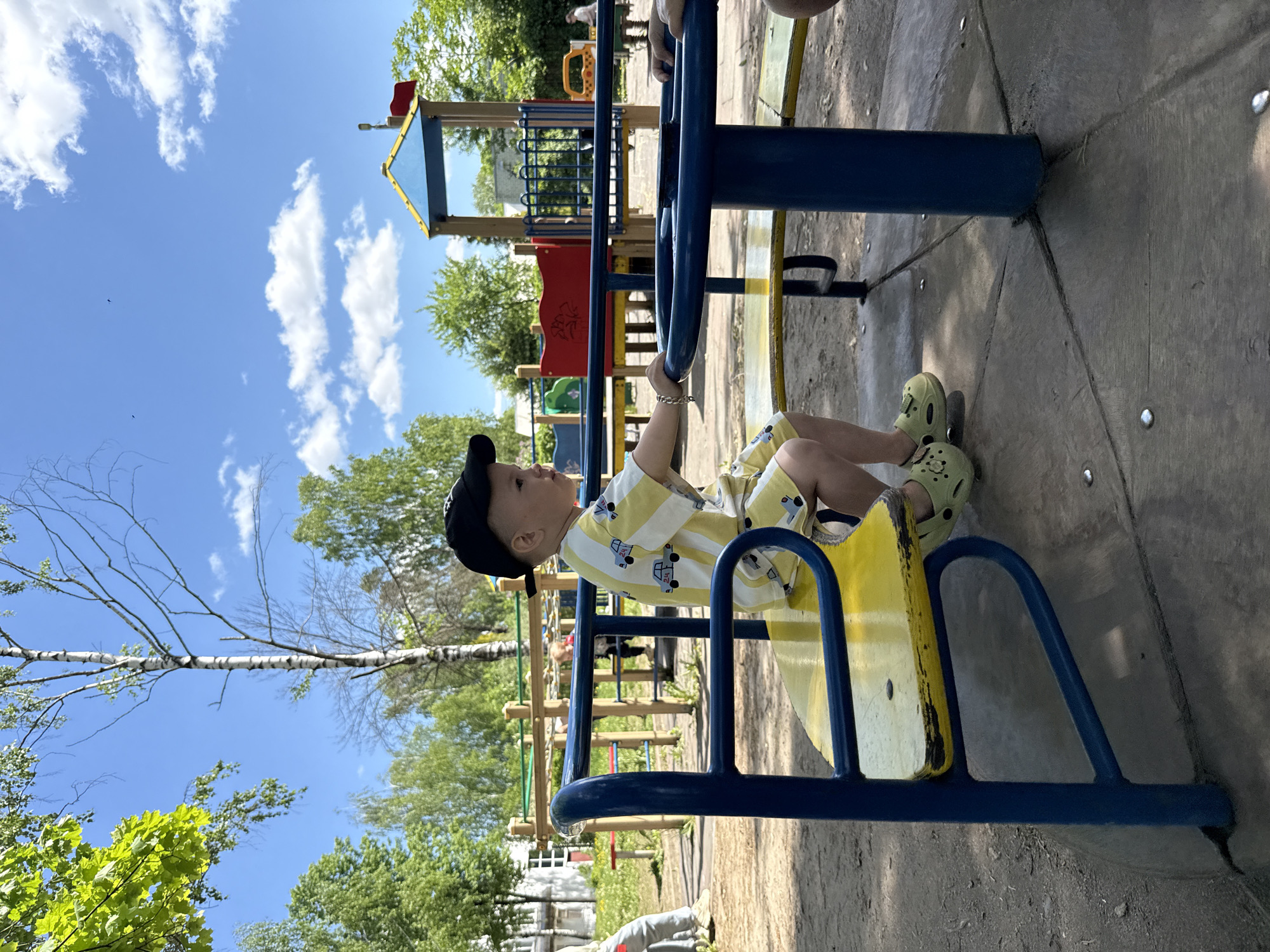 A young child, wearing a black baseball cap, a yellow and white striped shirt, yellow shorts with a car print, and green clogs, is sitting on a playground carousel. The carousel has a blue frame and a yellow seat. The child is holding onto the blue metal bar in front of them. The background shows other playground equipment with blue, red, and green elements, green trees, and a partly cloudy blue sky. The location appears to be a playground in a park or residential area. Rusanovo, Russia is the location specified.