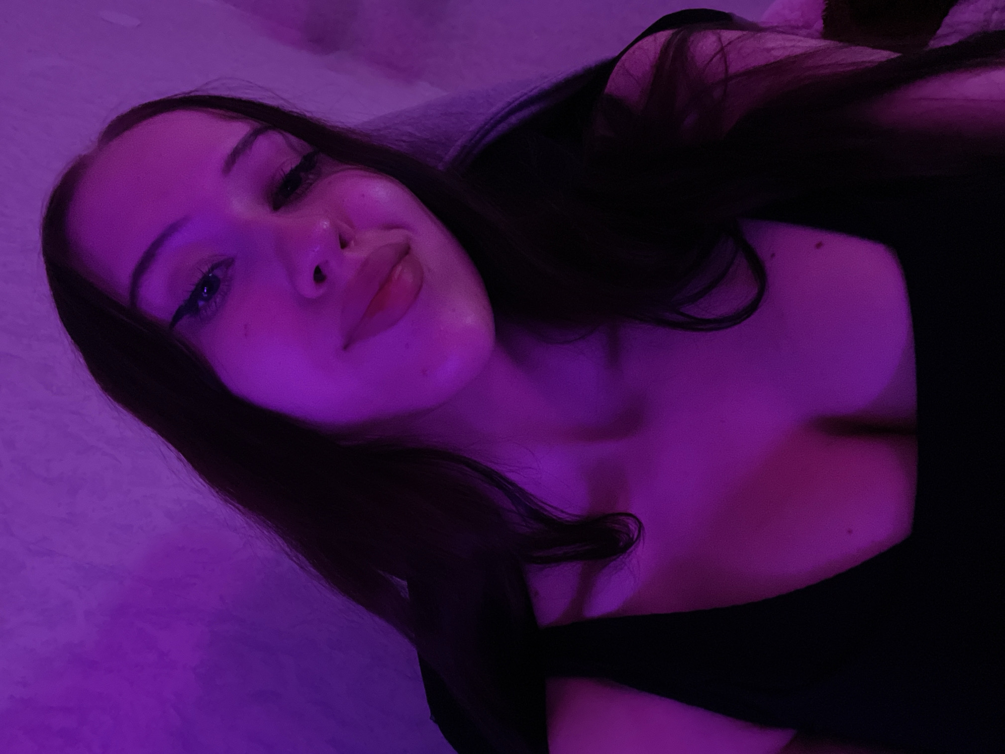 This is a close-up selfie of a young woman taken indoors. She has long, dark hair and is wearing a black top. The lighting is predominantly purple, creating a dramatic and moody atmosphere. Her face is angled towards the camera, and she appears to be looking downwards. Her makeup includes eyeliner and lip gloss. The background is out of focus and appears to be a wall with a textured surface. There is no indication of the specific location within Votkinsk, Russia, or any specific actions or interactions.
