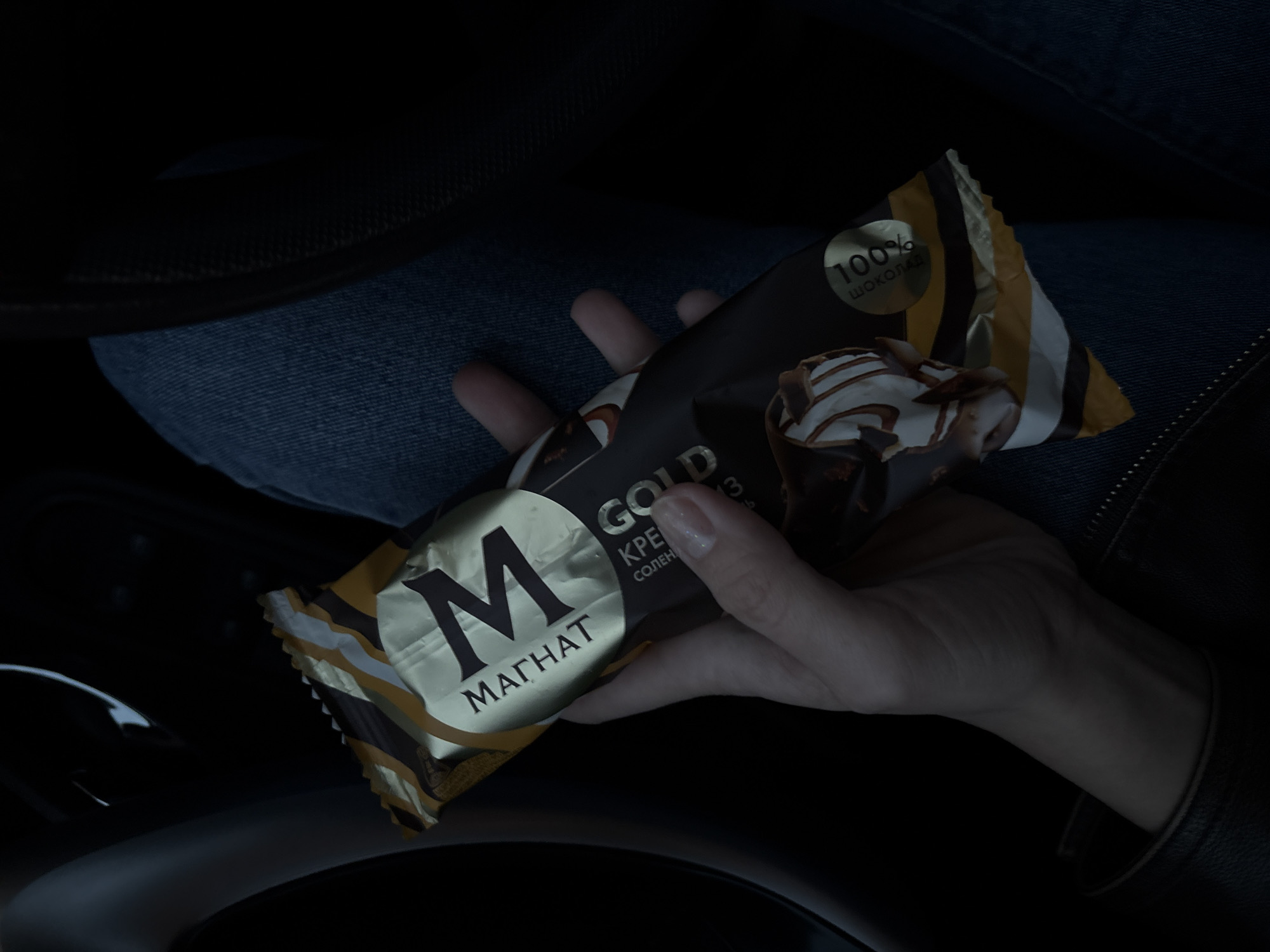 A hand holds a Magnum "Magnat" ice cream bar in a car. The packaging is primarily gold, black, and white with stripes. The brand "Magnat" is printed in large black letters in Cyrillic. The packaging also advertises "GOLD" flavor and "100% chocolate". The person is wearing jeans and a dark jacket. The location is likely indoors due to the darkness, but the setting is in the interior of a car. There is no indication that the location is Ust-Ilimsk, Russia.