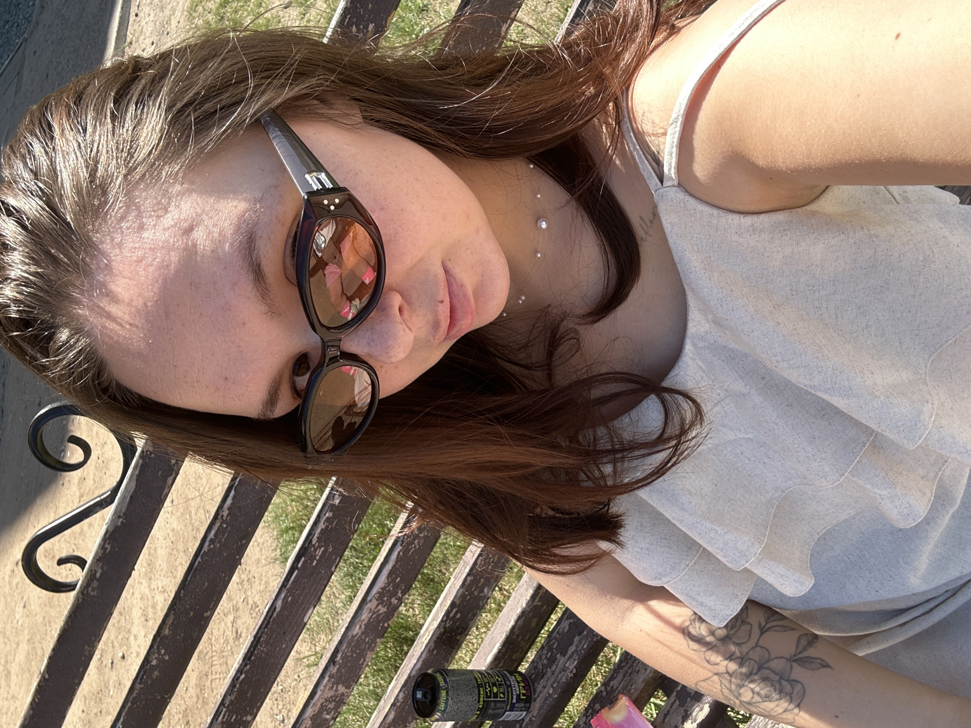 The image is a selfie of a woman lying on a park bench in Chaikovsky, Russia. She is wearing sunglasses and a light-colored, ruffled top. A beverage can and a pink object are also on the bench beside her. The bench is made of wood and metal, and there is grass visible in the background. The woman has a tattoo on her arm. She also appears to have a neck piercing.