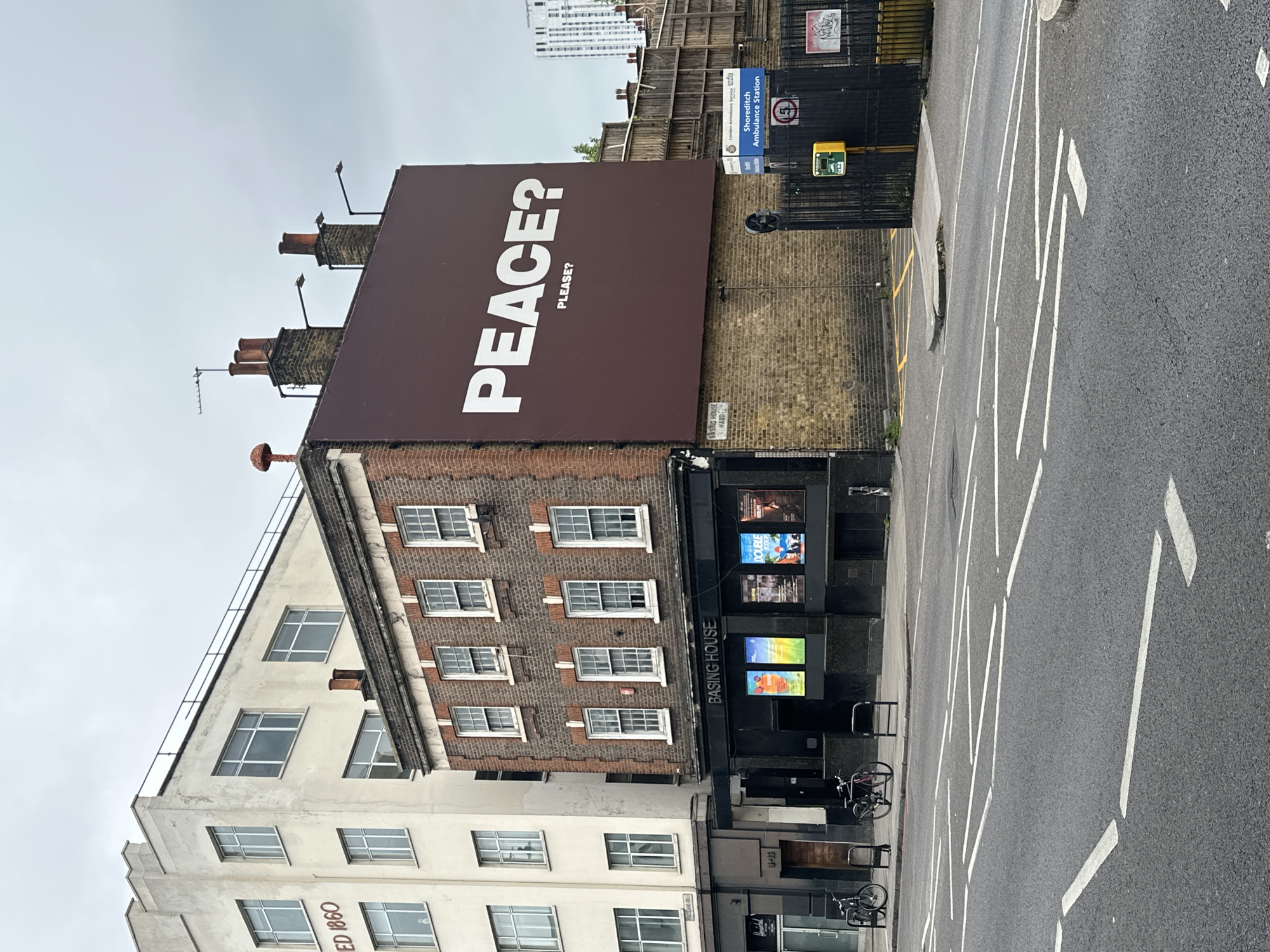 The image shows a building in Shoreditch, London, United Kingdom. The building, named "Basing House," has a prominent billboard on its roof that reads "PEACE? PLEASE?". The architectural style combines brick and white painted sections. A sign for the "Shoreditch Ambulance Station" is visible nearby. The street in front of the building has white painted lane markings. Several posters are visible in the windows of the ground floor. Bicycles are parked in front of the building's entrance. The day is overcast.