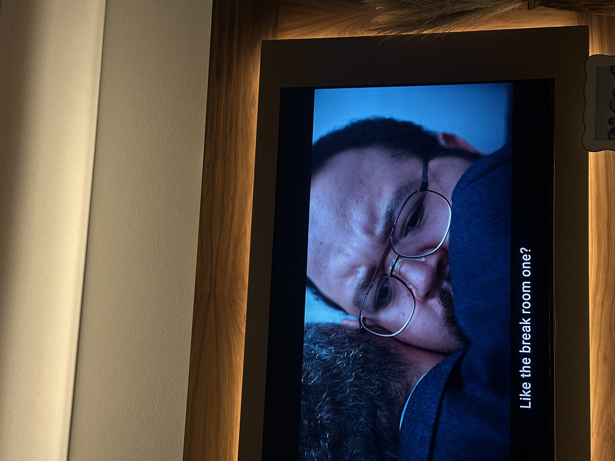 A television screen displays a scene of a man with glasses resting his head on another person's head with gray hair. The text "Like the break room one?" is visible on the screen. The television is surrounded by a wooden frame with a light fixture above the screen. The setting is indoors with a white wall on the left. The location is unknown.