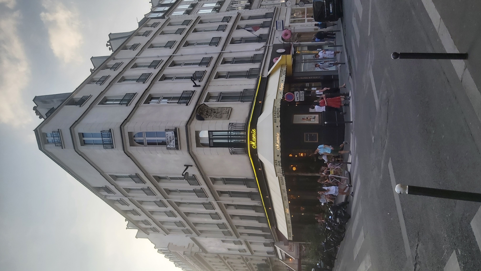 This is a street-level, full-frame, vertical, eye-level shot of a corner building in Paris, France, likely taken in the late afternoon or early evening as indicated by the lighting. The building is a multi-story structure with a classic Parisian architectural style, featuring cream-colored facades, numerous windows with black wrought-iron balconies, and mansard roofs with dormer windows.
The ground floor of the building is occupied by a restaurant or cafe named "Mamie," which is prominently displayed on a yellow awning with neon lighting. The awning is supported by poles and stretches across the storefront, with outdoor seating visible underneath. Several patrons are seated at tables, enjoying the ambiance. The facade of Mamie features dark windows and doorways, with smaller signs indicating "CAFE BAR" and "RESTAURANT BISTRO."
To the right of Mamie, there are other businesses with signage, including "POLICE" visible on a sign attached to the building. A pink donut-shaped decoration is also visible on the right side of the building, suggesting a bakery or dessert shop.
The street in the foreground is paved with asphalt and marked with pedestrian crosswalks. Several motorcycles are parked along the sidewalk to the left of the building. People are walking on the sidewalk and crossing the street, engaged in everyday activities. A dark-colored van is parked further down the street on the right. The sky above is overcast with scattered clouds. The overall scene depicts a lively urban environment in Paris.