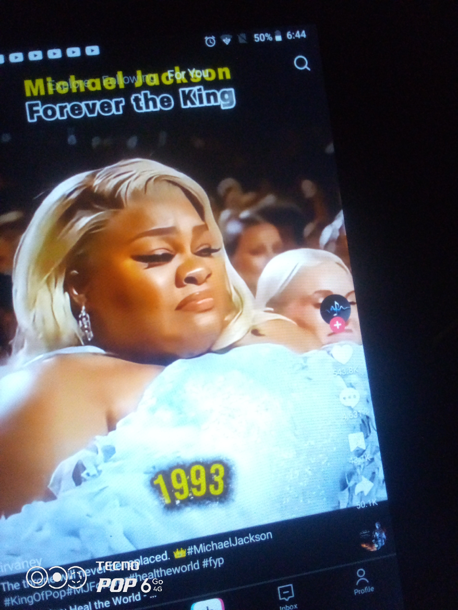 The image is a close-up, angled shot of a tablet screen displaying a video. The video appears to be a tribute to Michael Jackson, with text overlayed on the screen. The text reads "Michael Jackson Forever the King" in yellow and white fonts. A prominent yellow number "1993" is visible on the screen, likely indicating a year related to the video's content. The video itself shows a person with blonde hair in the foreground, with another person partially visible behind them. Social media interface elements such as like, comment, and share icons, along with user names and hashtags like "#KingOfPop" and "#MichaelJackson," are also present on the screen. The overall scene suggests a digital viewing experience of a tribute video on a personal device. The location in the city of Howayi, Nigeria, is not discernible from the visual information provided in the image.