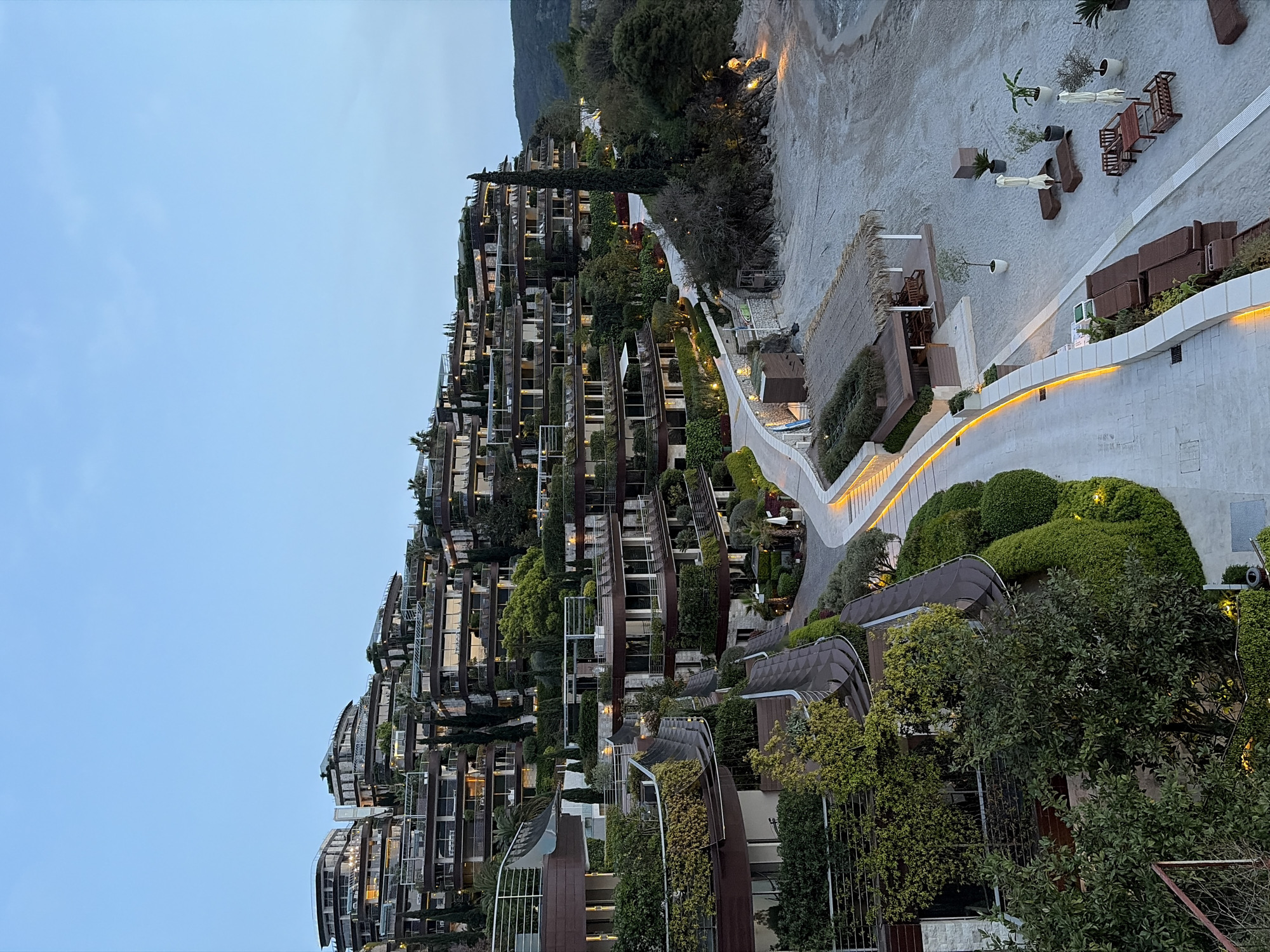 The image shows a multi-tiered residential complex built on a hillside, likely near a beach. The complex is characterized by many balconies and lush greenery interspersed among the units. A paved pathway with lighting leads down the slope towards the beach area, which features beach furniture and thatched structures. In the background, the sky is a pale blue. The hillside architecture and coastal setting suggest the location could be in the city of Budva, Montenegro, known for its similar geographical features and architectural styles.