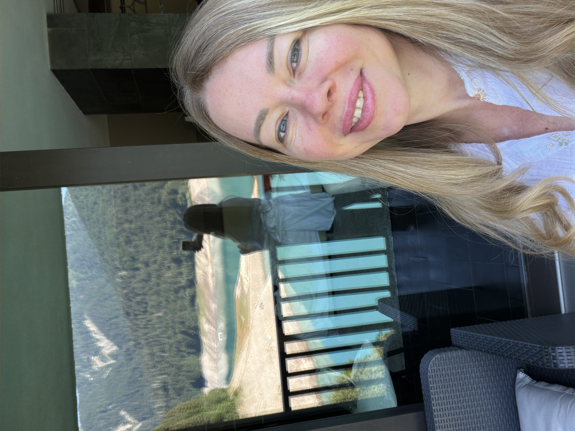 A woman with long blonde hair and blue eyes smiles at the camera. She is wearing a white blouse with floral embroidery. The background shows a balcony with a railing, a lake, and mountains covered in trees. A reflection in the glass shows another person holding a phone. There is outdoor furniture on the balcony. The scene is likely a hotel room or a residential balcony in Lateis/Latais, Italy, given the mountainous landscape and the presence of a lake.