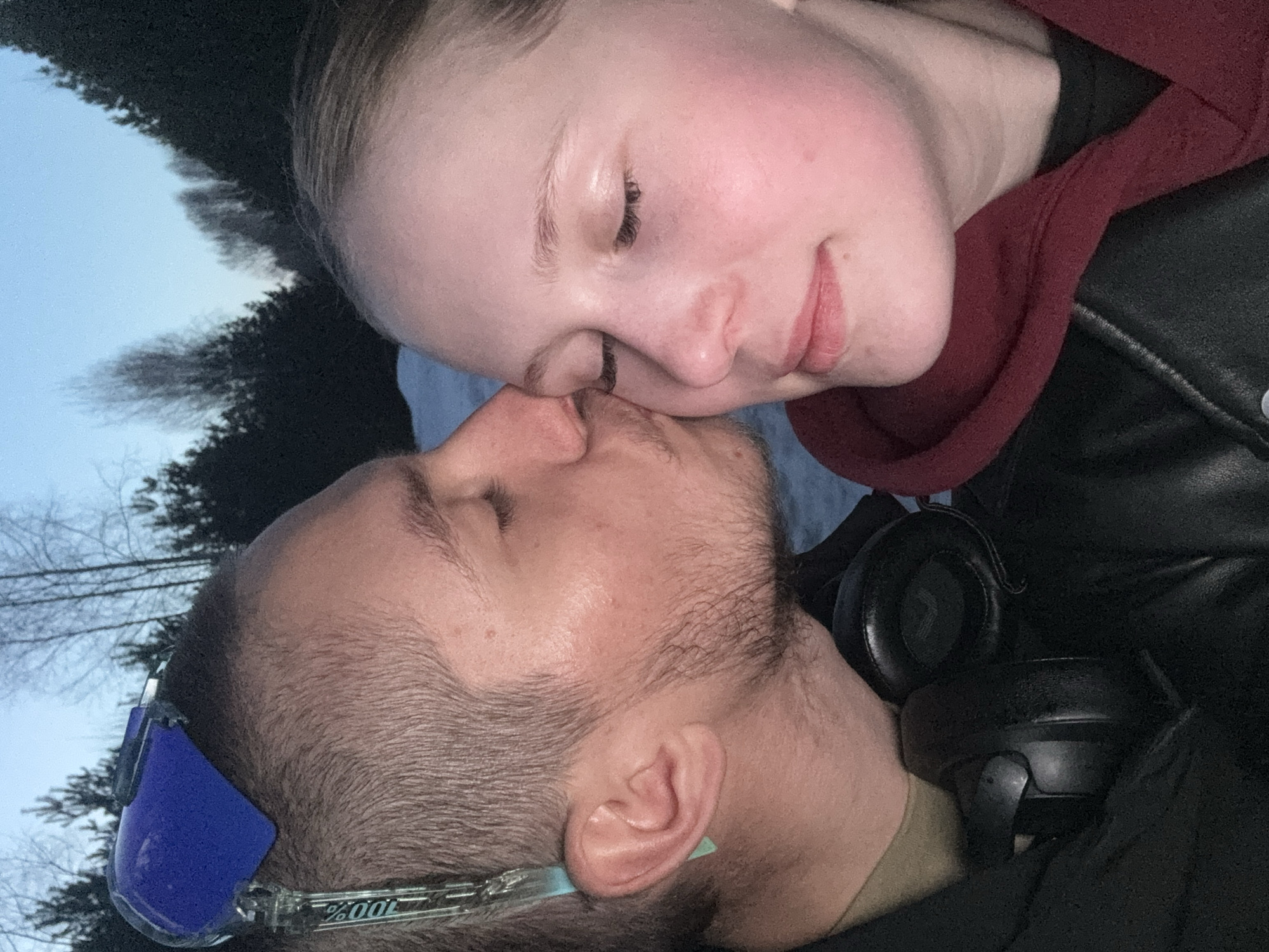 Two people are closely positioned, their faces touching. The person on top has fair skin, closed eyes, and a slight smile. They are wearing a maroon hoodie under a black jacket. The person below has short hair, stubble, closed eyes, and is wearing sunglasses. Black headphones are visible next to their head. In the background, there are trees and a muted sky. There is no evidence to suggest the image was taken in the city of Chaikovsky, Russia.