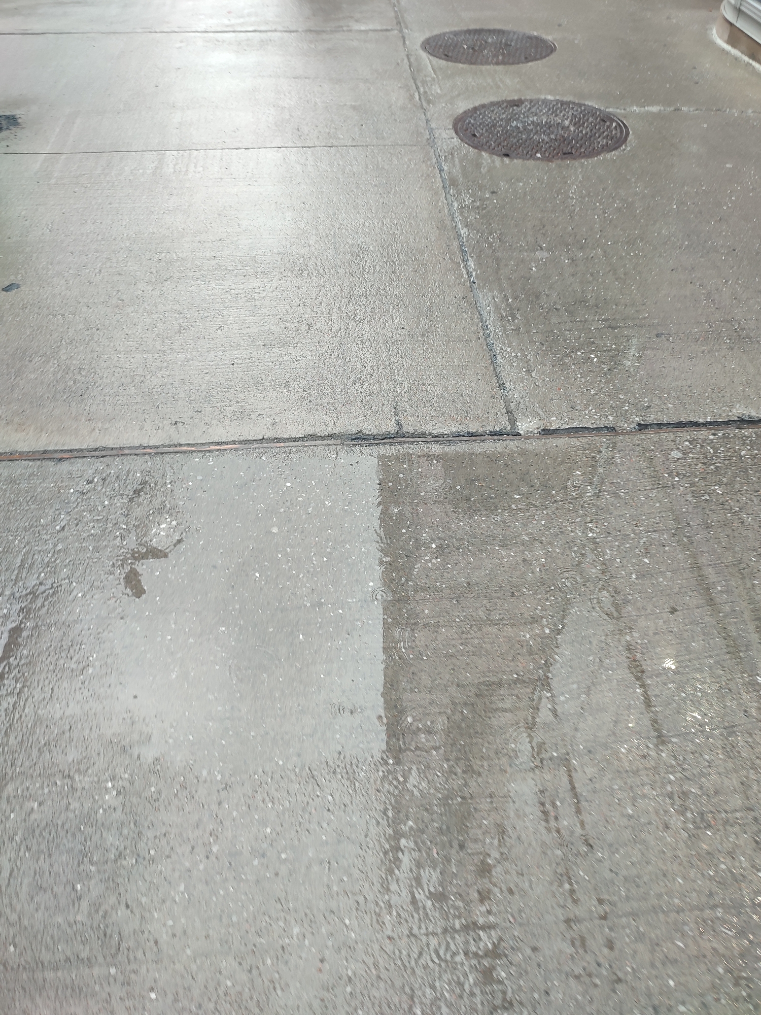 The image shows a wet concrete surface, likely a sidewalk or a paved area, with two circular manhole covers visible in the upper right portion. The concrete is divided by expansion joints, creating large rectangular sections. A significant portion of the lower left of the image is covered by a shallow puddle of water, reflecting the overcast sky. The wetness creates a darker, more reflective sheen on the concrete compared to the drier areas. The surface texture is granular, typical of concrete with exposed aggregate. There are no people or animals visible in the image. The scene suggests a recent rainfall or a damp environment. The lighting is diffused, indicating it is likely daytime with overcast weather conditions. The presence of manhole covers suggests an urban or suburban setting, possibly a street or a public area. No visible text is present in the image. The overall mood is neutral, focusing on the texture and reflectivity of the wet concrete surface. The setting is outdoors.