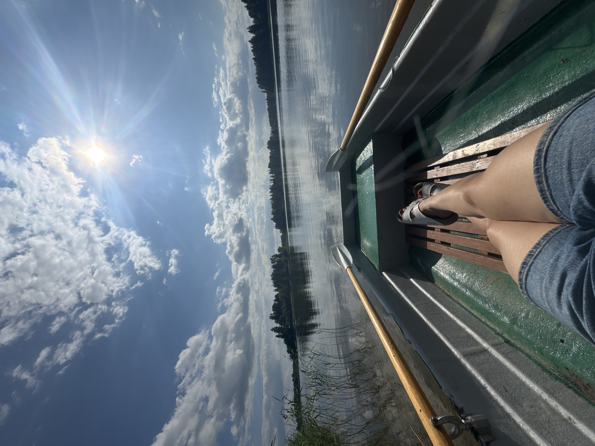 This media file depicts a person's legs and feet, wearing denim shorts and white sandals, resting inside a rowboat. The boat appears to be on a calm lake, with a long, tree-lined shore visible in the distance. The water reflects the sky and the trees. A wooden oar with a white blade rests in the boat, positioned for rowing. The sky is bright blue with fluffy white clouds and the sun is shining intensely, creating a lens flare effect and casting strong shadows. The overall impression is one of a sunny day spent on the water. The location is identified as Lendži, Latvia. No visible text is present.