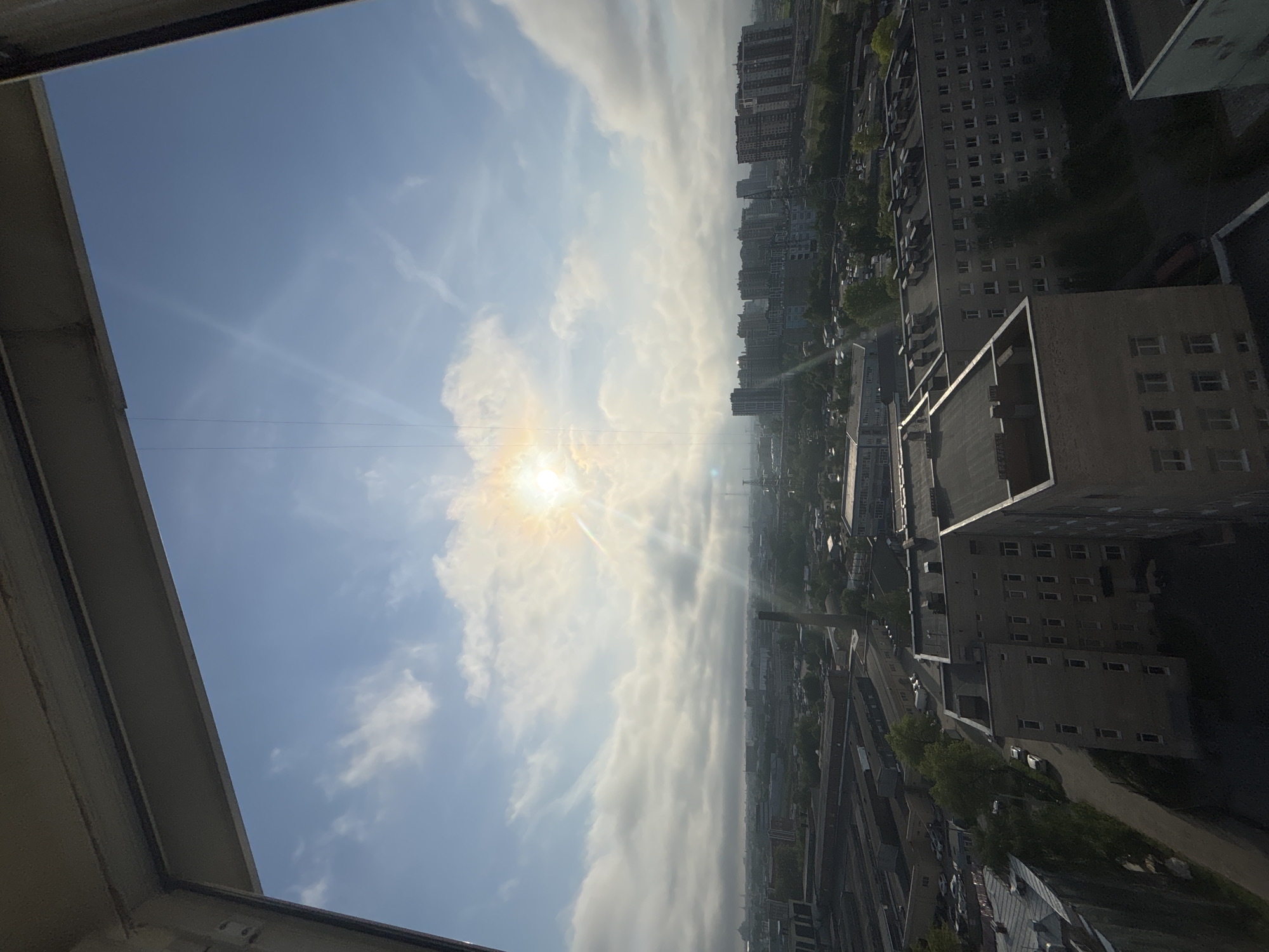 The image shows a cityscape from an elevated vantage point, possibly a high-rise building or apartment. The sky is visible with sunlight shining through a layer of clouds. Buildings of various heights are visible, suggesting an urban or suburban area. Roads and vegetation can be seen between the buildings. The overall environment appears to be a densely populated area.