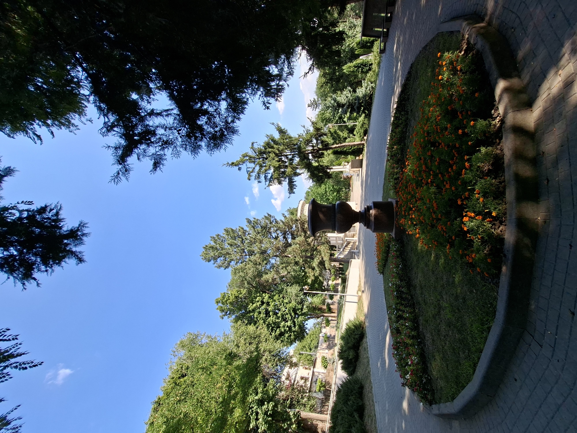 The image depicts a circular flowerbed in a park setting, likely in Chișinău, Moldova. The flowerbed is planted with vibrant orange marigolds and white and red flowers, arranged around a large, dark-colored urn situated at its center. The flowerbed is bordered by a curved stone edge and is surrounded by a paved pathway made of grey interlocking bricks, laid in a circular pattern.
The pathway curves around the flowerbed, leading into the wider park area. In the background, lush green trees of various species form a dense canopy, creating a dappled light effect on the path and ground. Sunlight filters through the leaves, casting shadows. Further in the distance, there are more trees and some architectural elements, possibly part of a building or monument, visible on the left. The sky is a clear blue with a few wispy white clouds. The overall scene suggests a well-maintained public park, possibly a garden or a promenade area, during a sunny day.