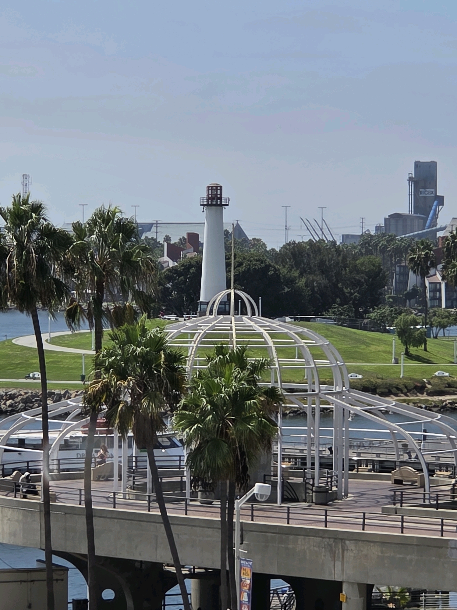 The image shows a view of Rainbow Harbor in Long Beach, California, United States of America. In the foreground, a concrete pedestrian bridge with a white, arched, open-air structure extends over the water. Several palm trees are visible in front of and on the bridge. On the bridge, there are benches and a few people. Beyond the bridge, a body of water, likely part of the harbor, is visible. Further back, a grassy hill leads up to a white lighthouse with a red roof. Beyond the lighthouse, there are various buildings, trees, and industrial structures, including a tall, blue and grey industrial silo. The sky is overcast.