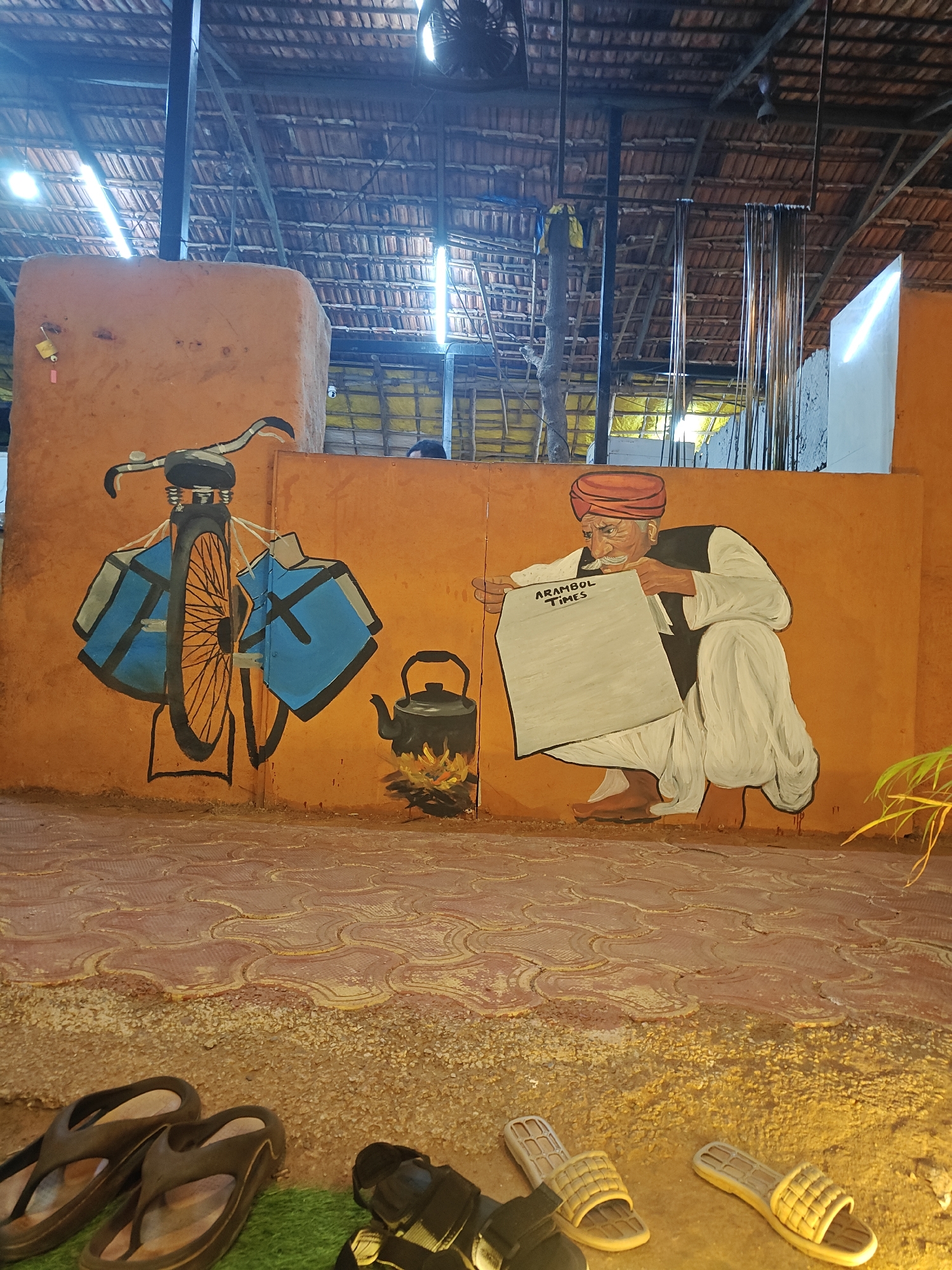 The image shows an outdoor area with a mural painted on an orange wall. The mural depicts a bicycle with blue panniers, a kettle on a fire, and a man in traditional Indian clothing squatting and reading a newspaper titled "ARAMBOL TIMES". In the foreground, there are several pairs of sandals. The ground is paved with reddish-brown patterned tiles and loose gravel. The ceiling above is made of woven thatch or similar material. The location appears to be in a city of Unknown, Unknown.