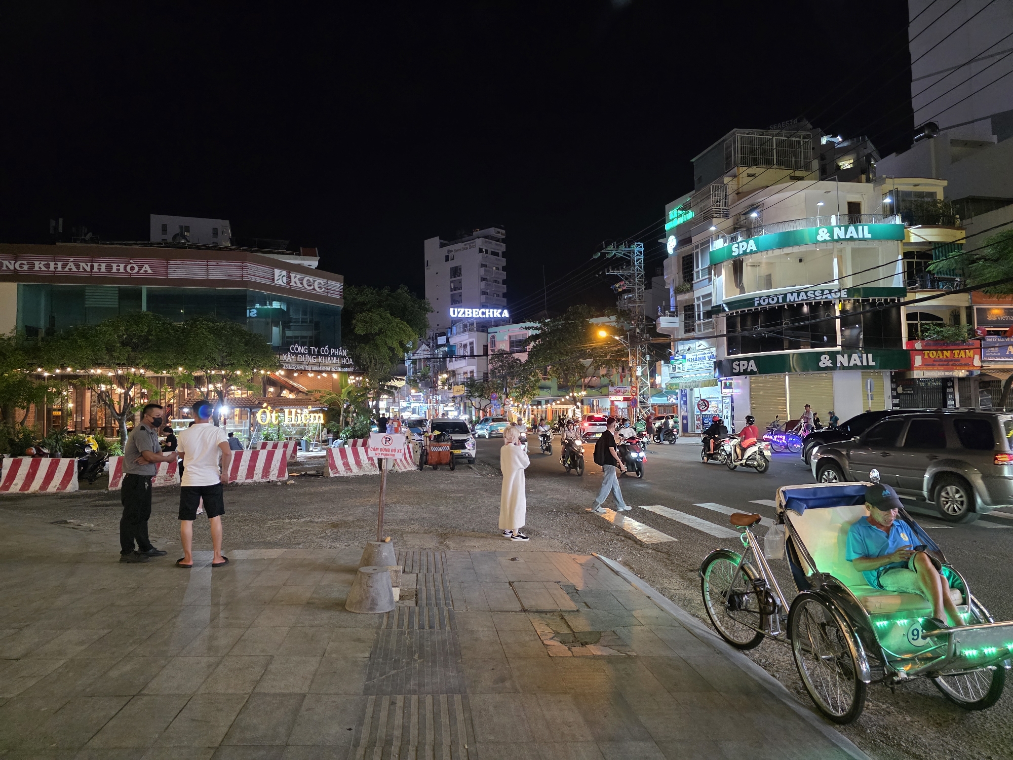 This media file presents a vibrant night street scene in Nha Trang, Vietnam, illuminated by numerous artificial lights against a dark sky.
In the foreground on the left, two men are standing on a sidewalk paved with large grey tiles. One man is dressed in a dark security uniform with a face mask, while the other wears a white t-shirt and dark shorts. Behind them are red and white striped barriers, and string lights adorn trees and an outdoor dining area. A white sign next to the barriers explicitly states "CẤM DỪNG ĐỖ XE" (No Stopping Parking) and "NO PARKING."
Further down the street, various people are visible, including a person in a long white robe standing near a pedestrian crossing. The street is busy with traffic, featuring cars, numerous motorcycles, and scooters.
On the right side of the road, a green LED-lit cyclo (pedicab) is parked near the curb. A man in a blue shirt and cap is seated comfortably inside the cyclo, engrossed in his phone. The number "9" is visible on the side of the cyclo.
Buildings line both sides of the street, brightly lit with prominent signs. On the left, a building displays "NG KHANH HOA" and "CÔNG TY CỔ PHẦN XÂY DỰNG KHÁNH HÒA" (Khanh Hoa Construction Joint Stock Company) along with "KCC". In the middle background, another building features "Seven Seas Hotel" and "UZBECHKA". To the right, a multi-story building prominently advertises "SPA & NAIL" and "FOOT MASSAGE" at different levels, with "AESTA" also visible. Other text includes "Ot Hiệm" (likely "Ốc Hiếm" meaning rare snails, referring to a restaurant) in the background, and "TOAN TAM", "CHUYỂN ĐIỆN THOẠI SI", "KHÁNH V", "Vietna", and "ĐẠI LÝ BẢN VỀ M" on other storefronts. The atmosphere is that of a bustling urban night, with commercial activity and evening traffic.